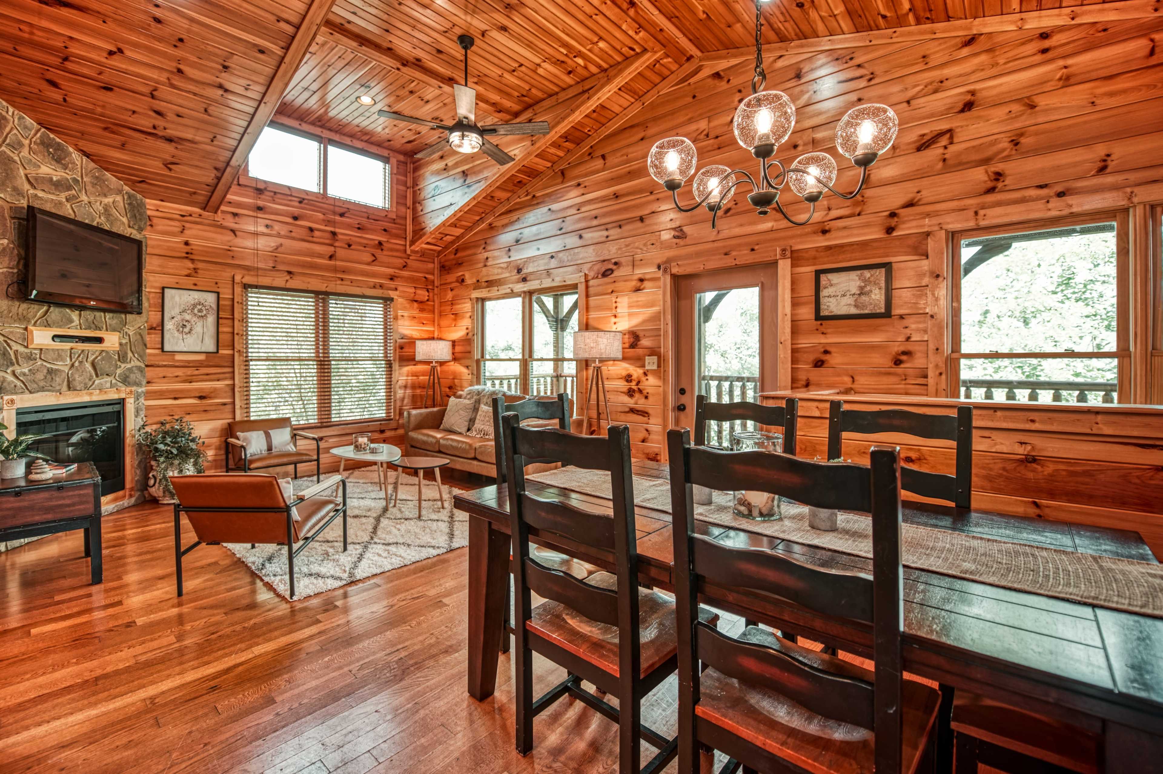 The interior of a wooden cabin features a dining table with black chairs, a stone fireplace, and large windows that allow natural light to fill the open living space.