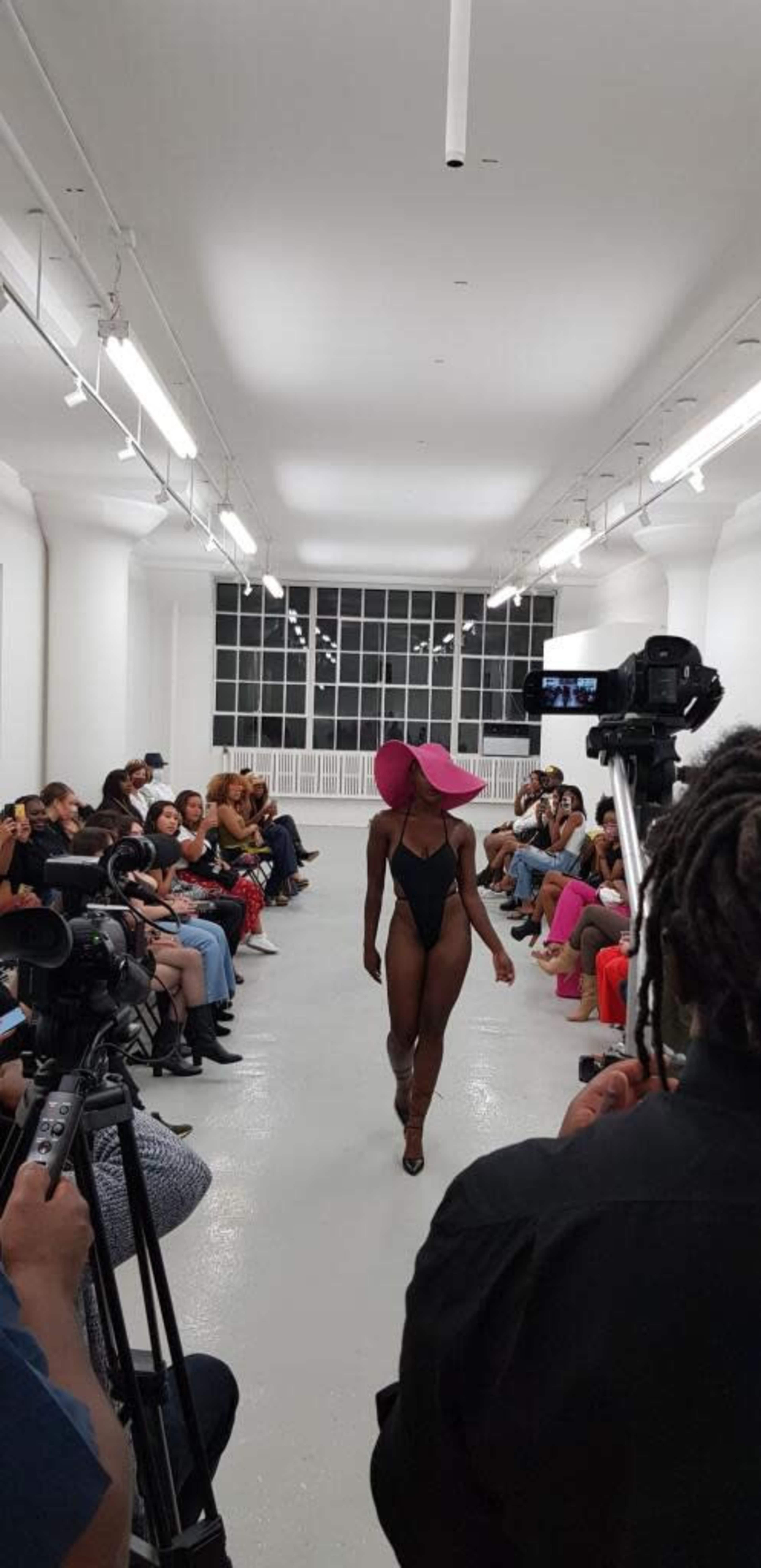A model walks down a runway in a swimsuit and large pink hat while photographers and an audience observe from the sides.