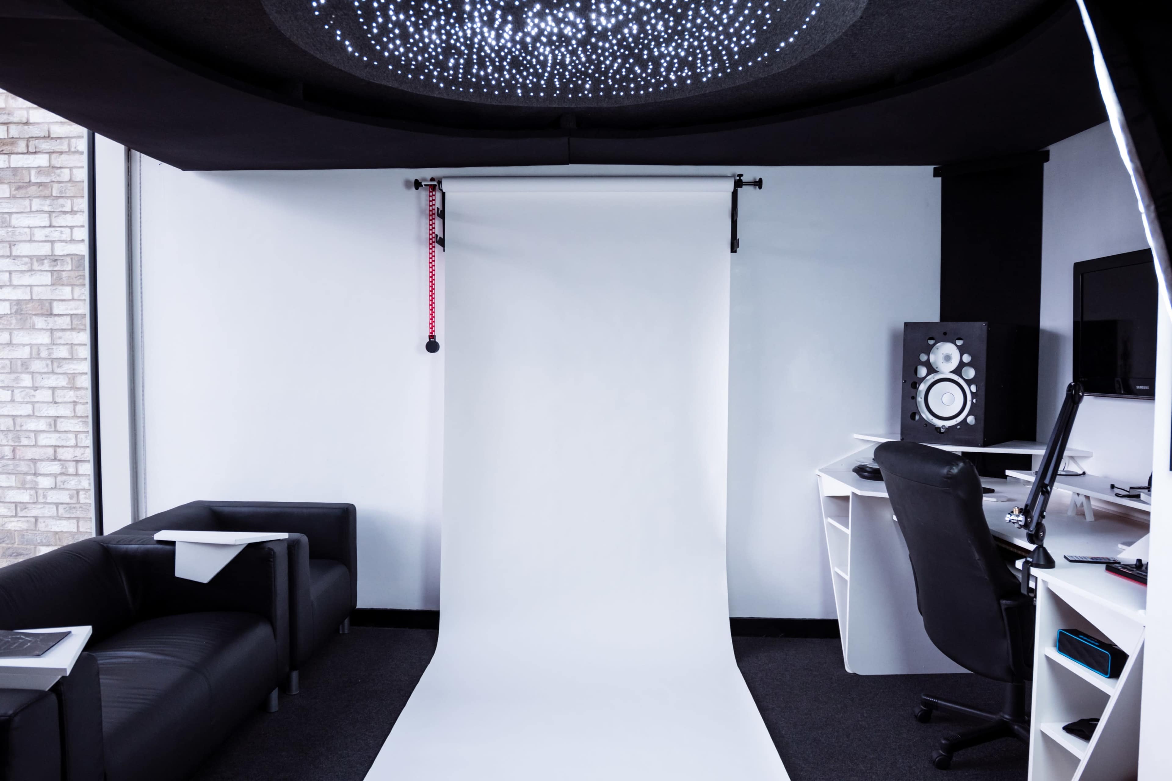 A minimalist studio space features a white backdrop, a black couch, and a desk with audio equipment and a computer.