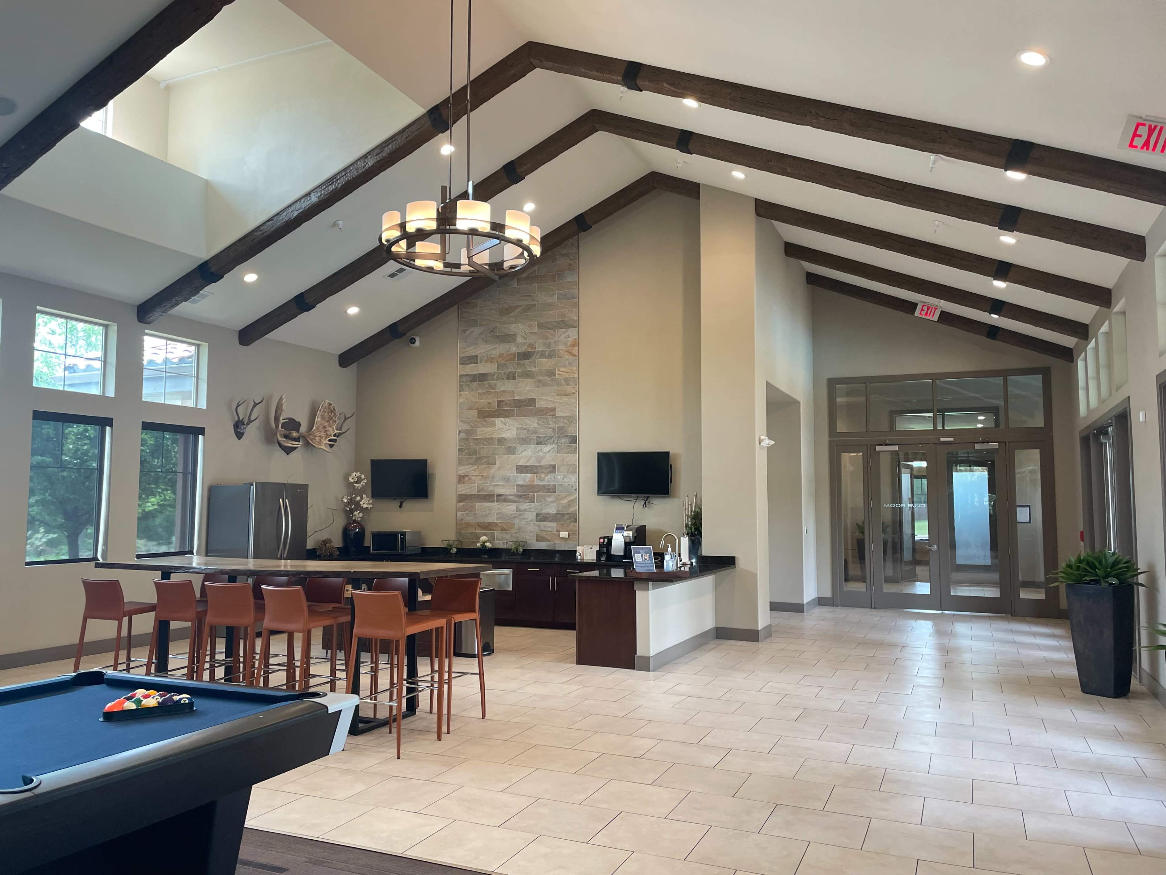 The image shows a spacious lobby area featuring a kitchen bar, several seating areas, and large windows with a stone accent wall and wooden beams overhead.