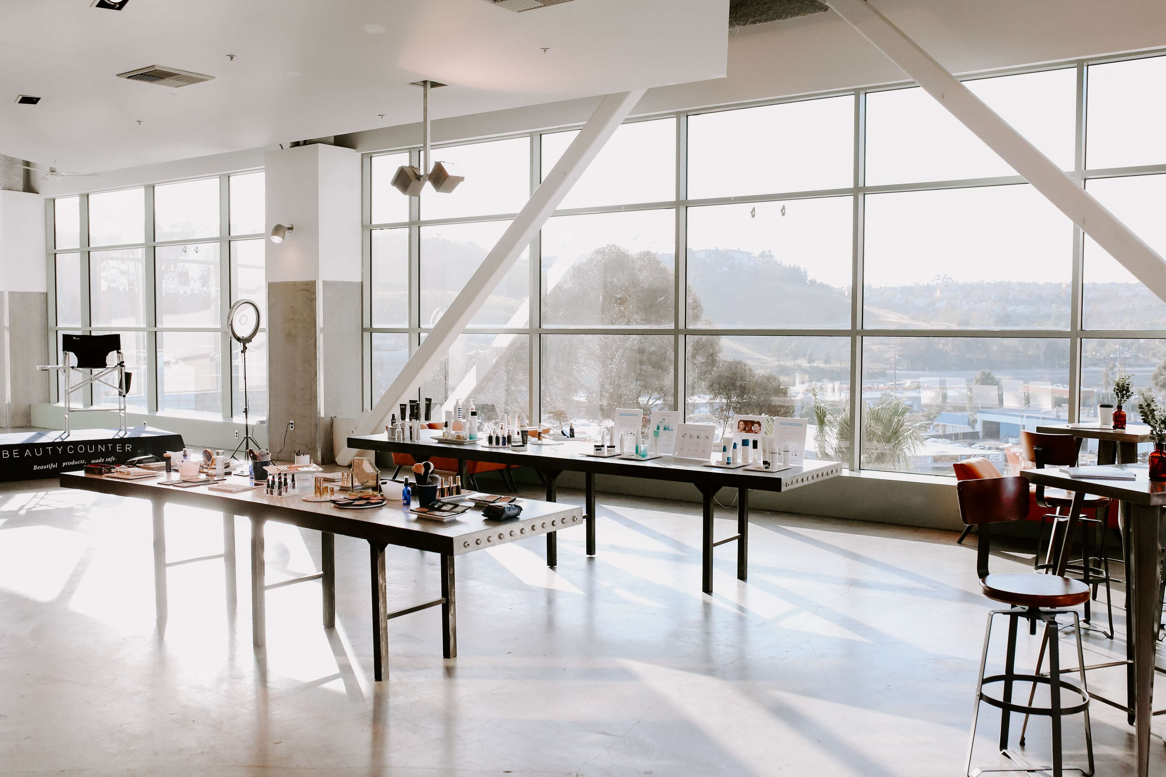 A spacious room with large windows, featuring several tables set up with various beauty products and a few chairs.