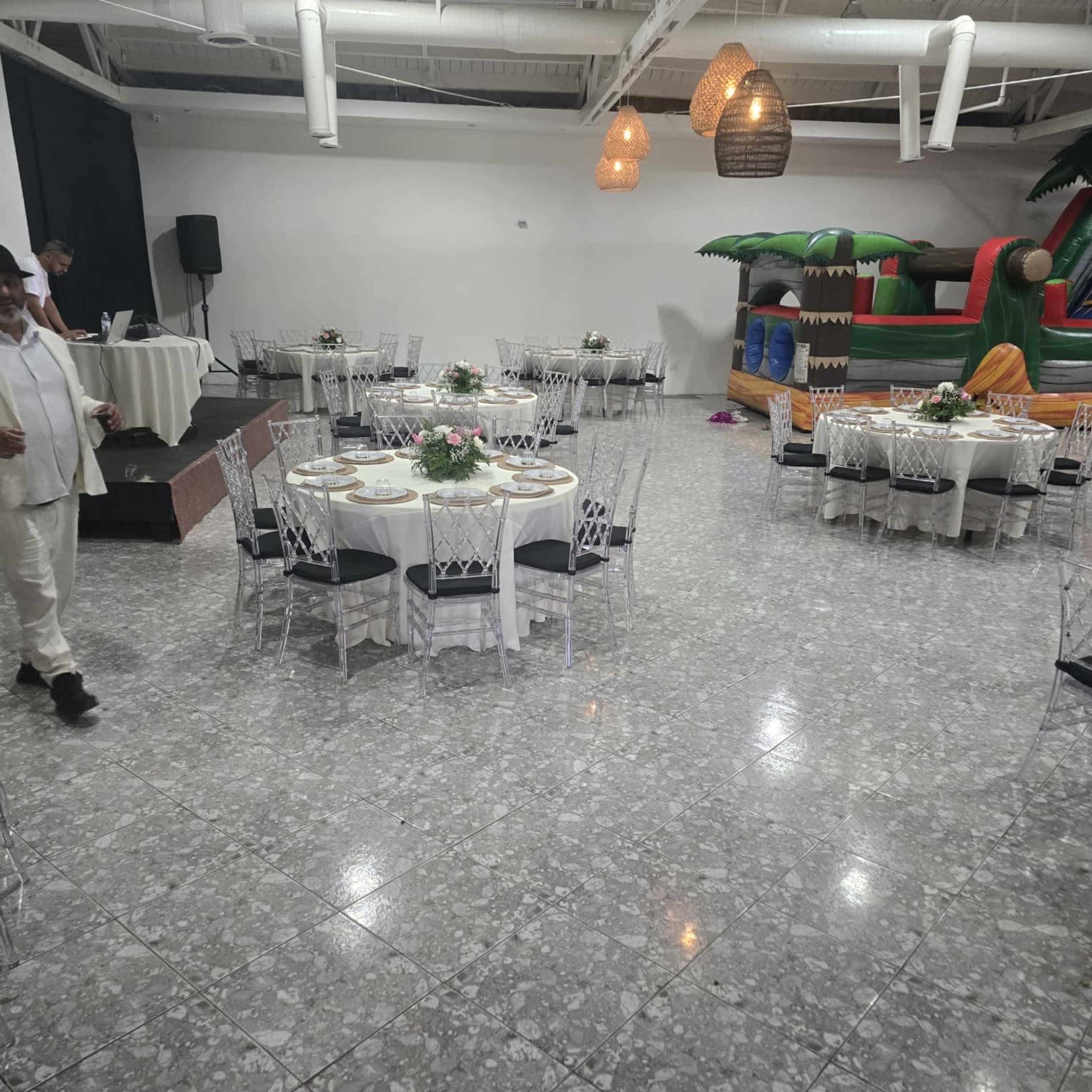The image shows an event space with round tables set for a gathering, decorated with floral centerpieces, and a bouncy castle in the corner.