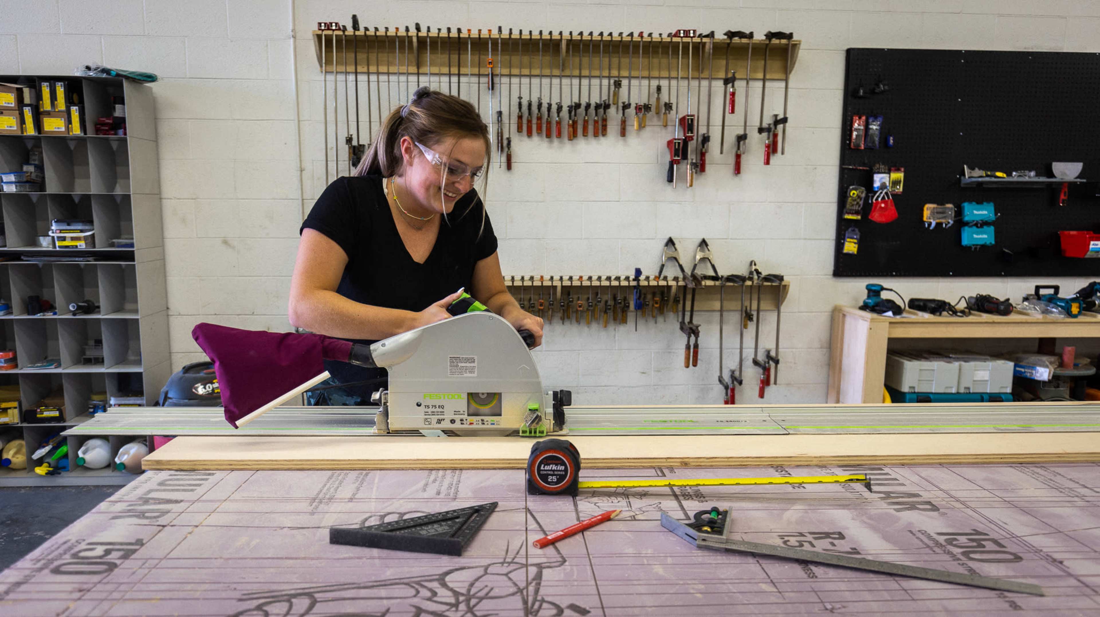 A woman operates a circular saw on a workbench in a well-organized workshop filled with various tools.