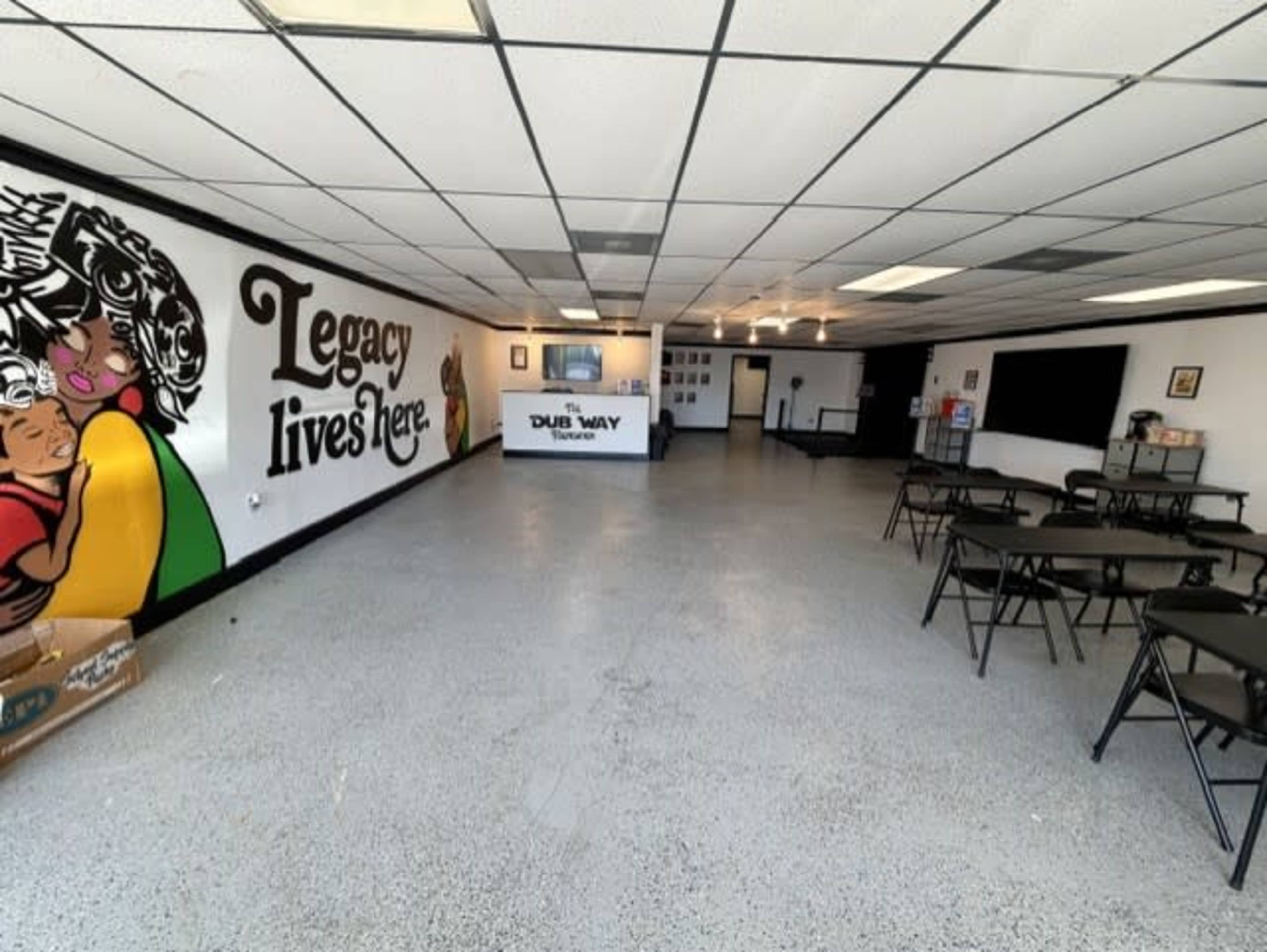 The image shows an empty community space with chairs and tables arranged, featuring a mural on the wall that reads "Legacy lives here."