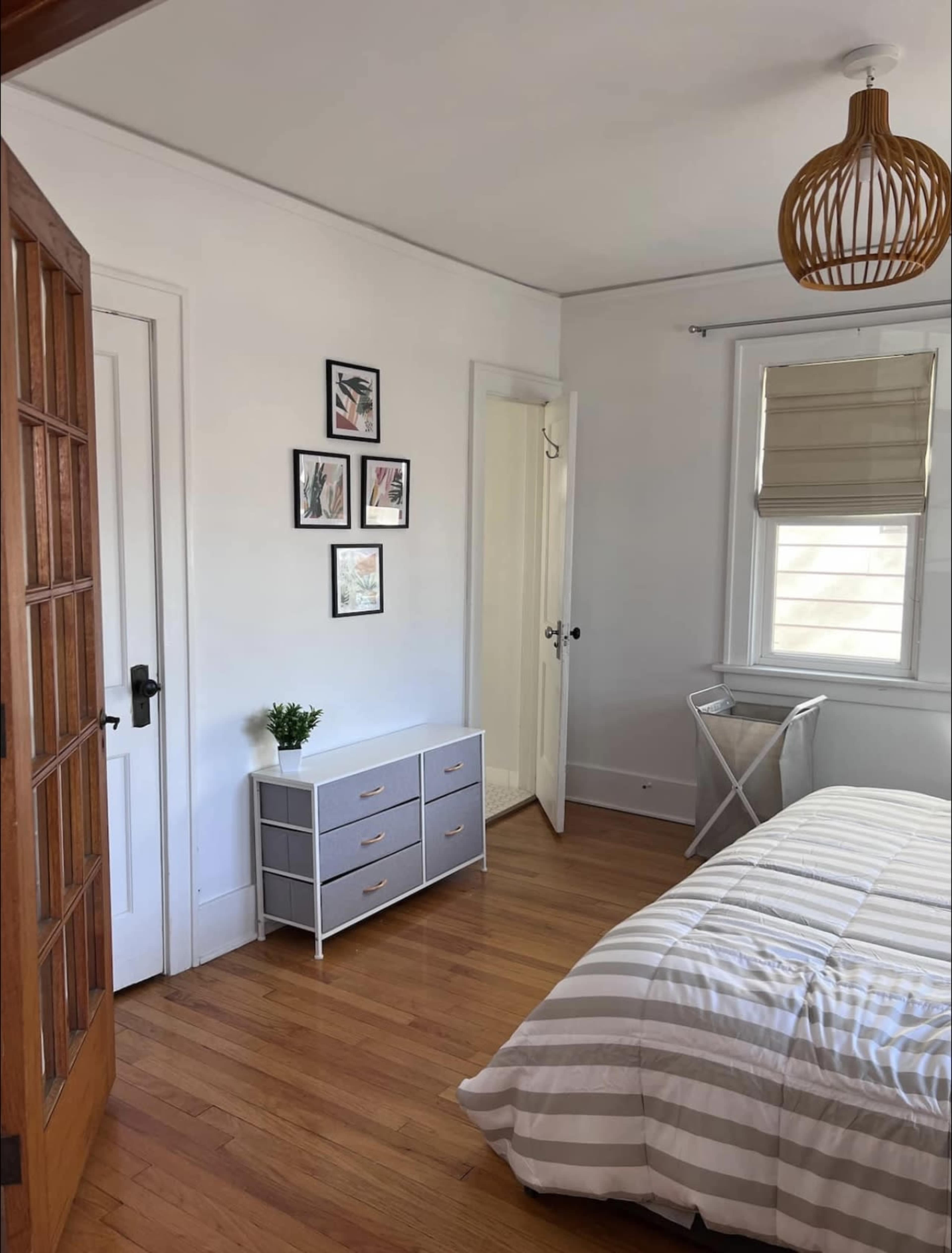 A room with wooden flooring, a bed with striped bedding, a gray dresser, framed pictures on the wall, and a door leading to another space.