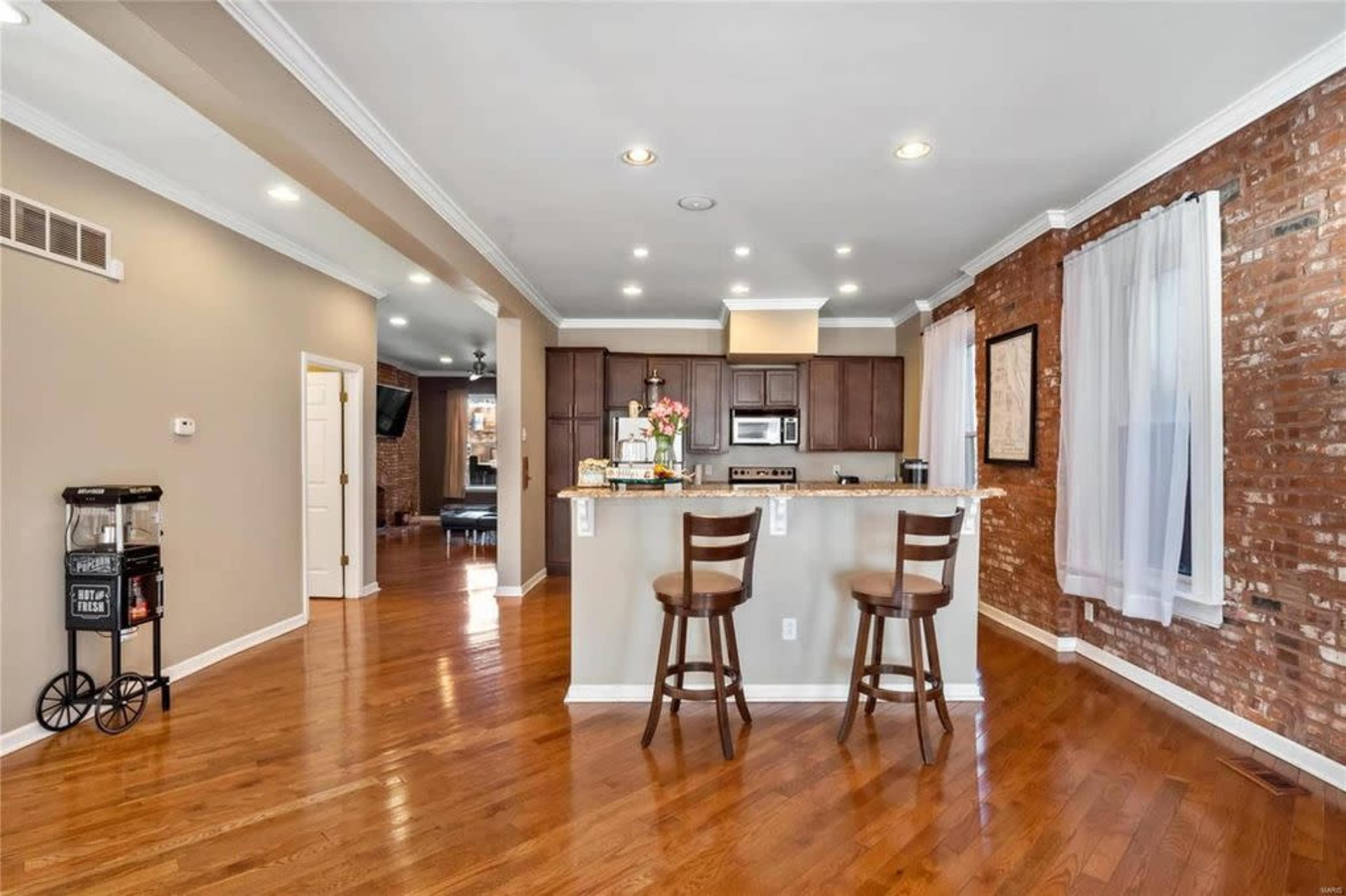 A kitchen area with bar stools, granite countertops, and wooden cabinets, adjacent to a living space with exposed brick walls and large windows.