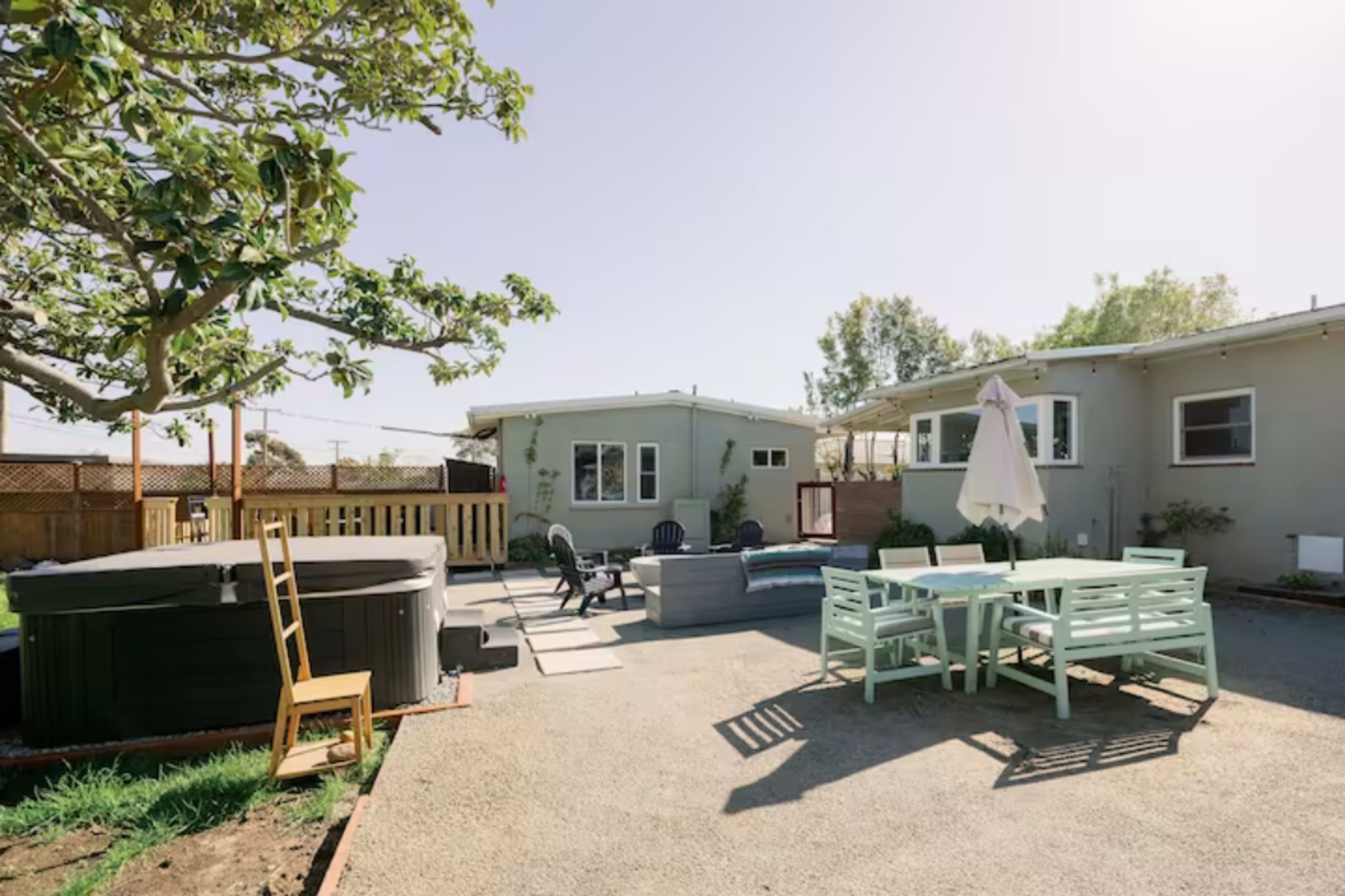 The image shows a spacious outdoor area with a hot tub, seating arrangements, and a dining table surrounded by a gravel surface and several trees.