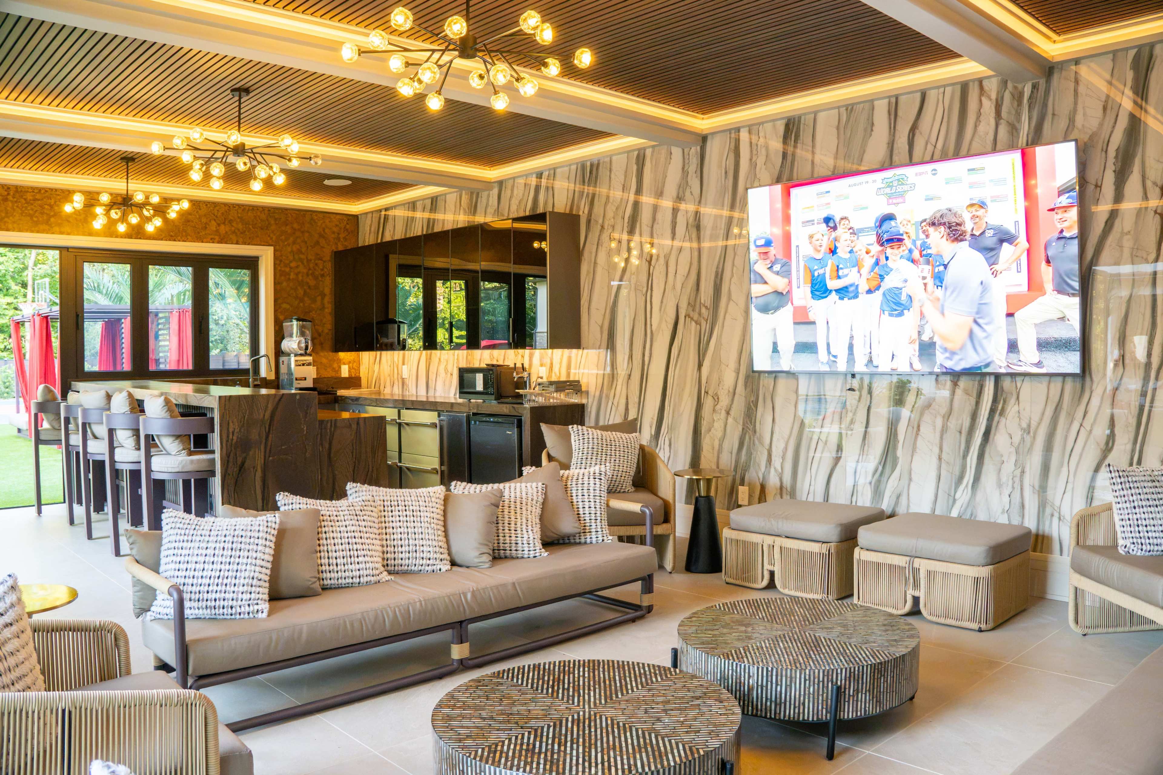 The image shows a modern lounge area with a large television, stylish seating, and a sleek kitchen backdrop featuring marble walls and warm lighting.