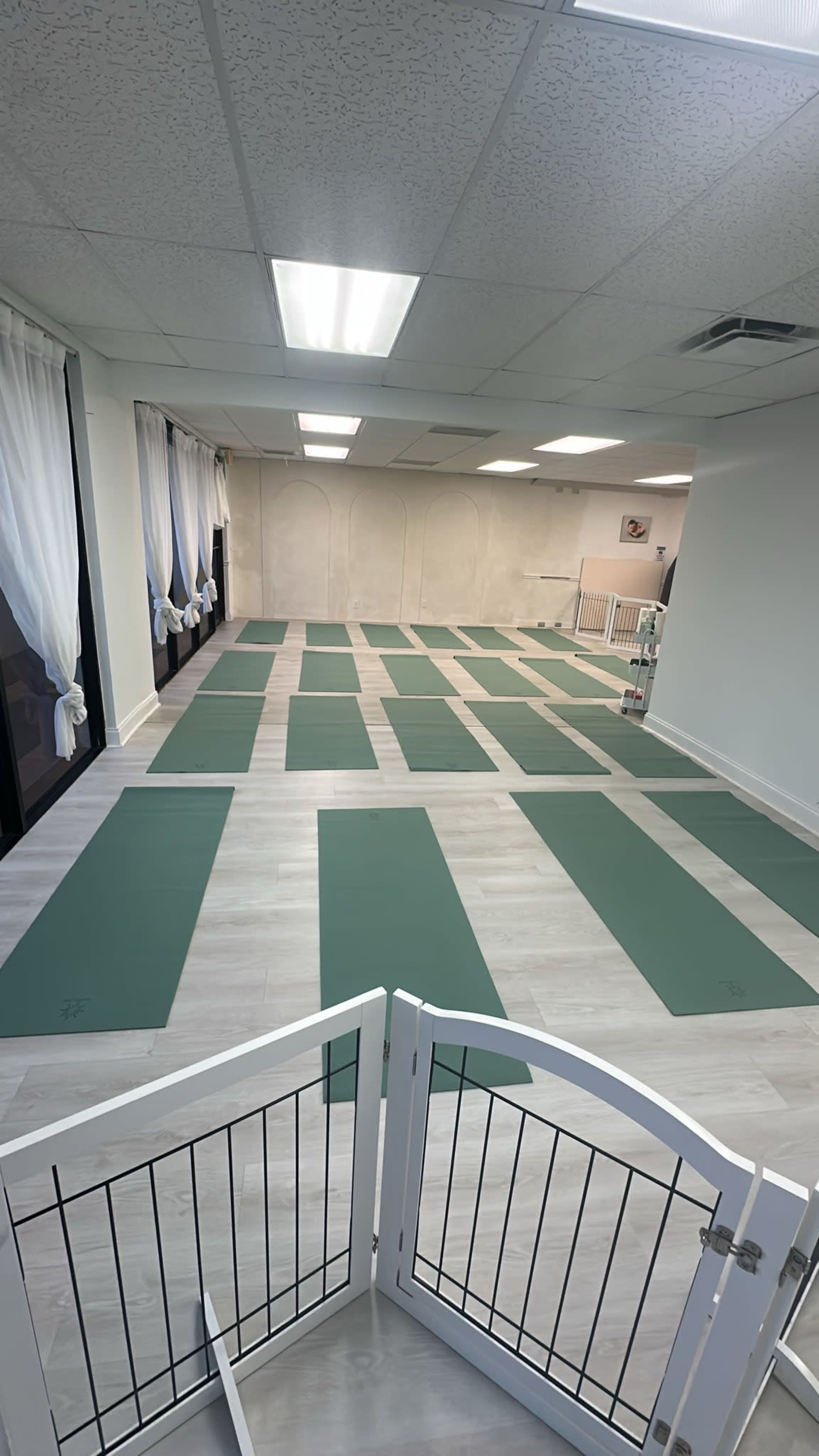 The image shows a spacious yoga studio with multiple green mats arranged neatly on a light-colored wooden floor.