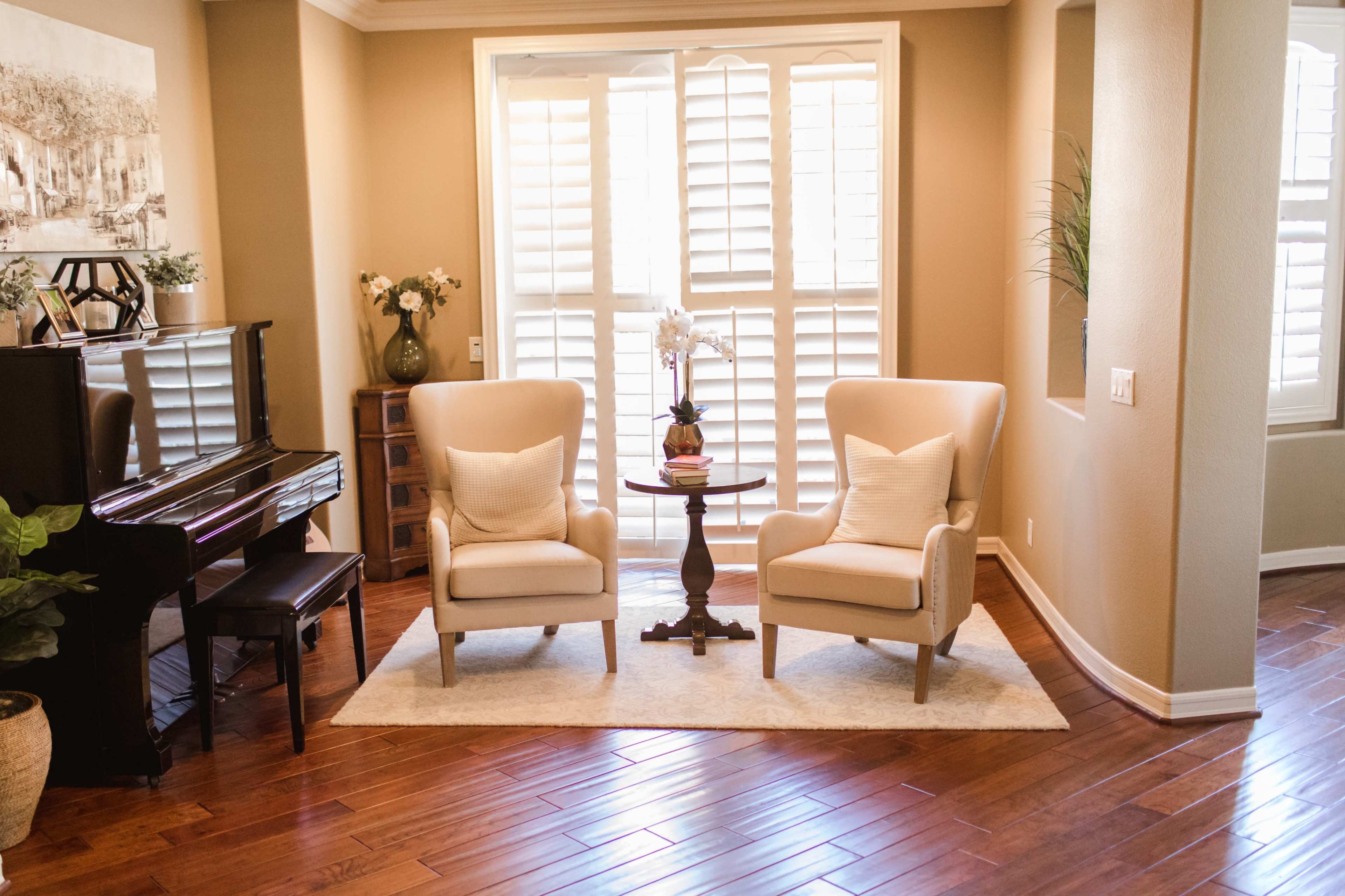 The image shows a cozy sitting area with two armchairs facing a small table, surrounded by natural light from a set of shutters, and a piano nearby.