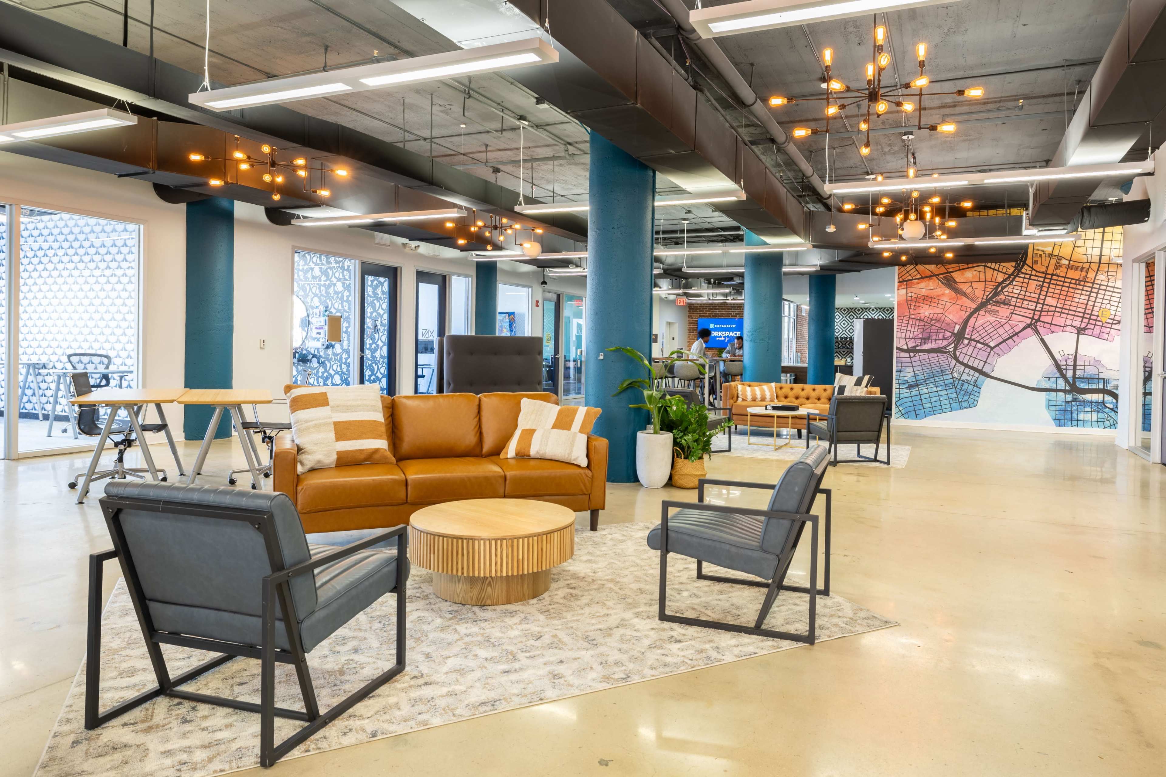 The image shows a modern coworking space with a mix of seating arrangements, a central orange sofa, and large windows letting in natural light.