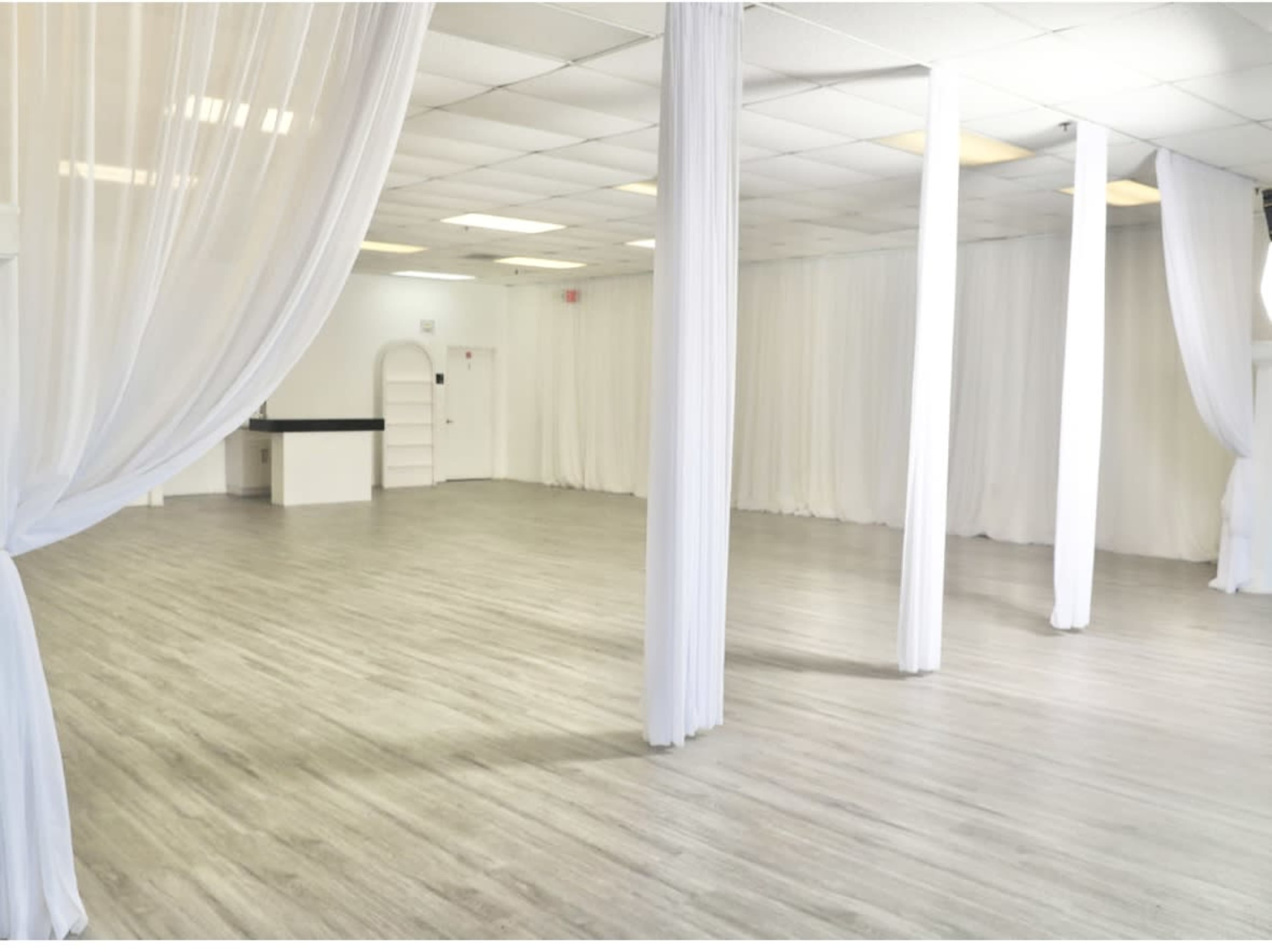 The image shows a spacious interior with light wood flooring, white drapery on pillars, and softly lit overhead lights in an empty room.