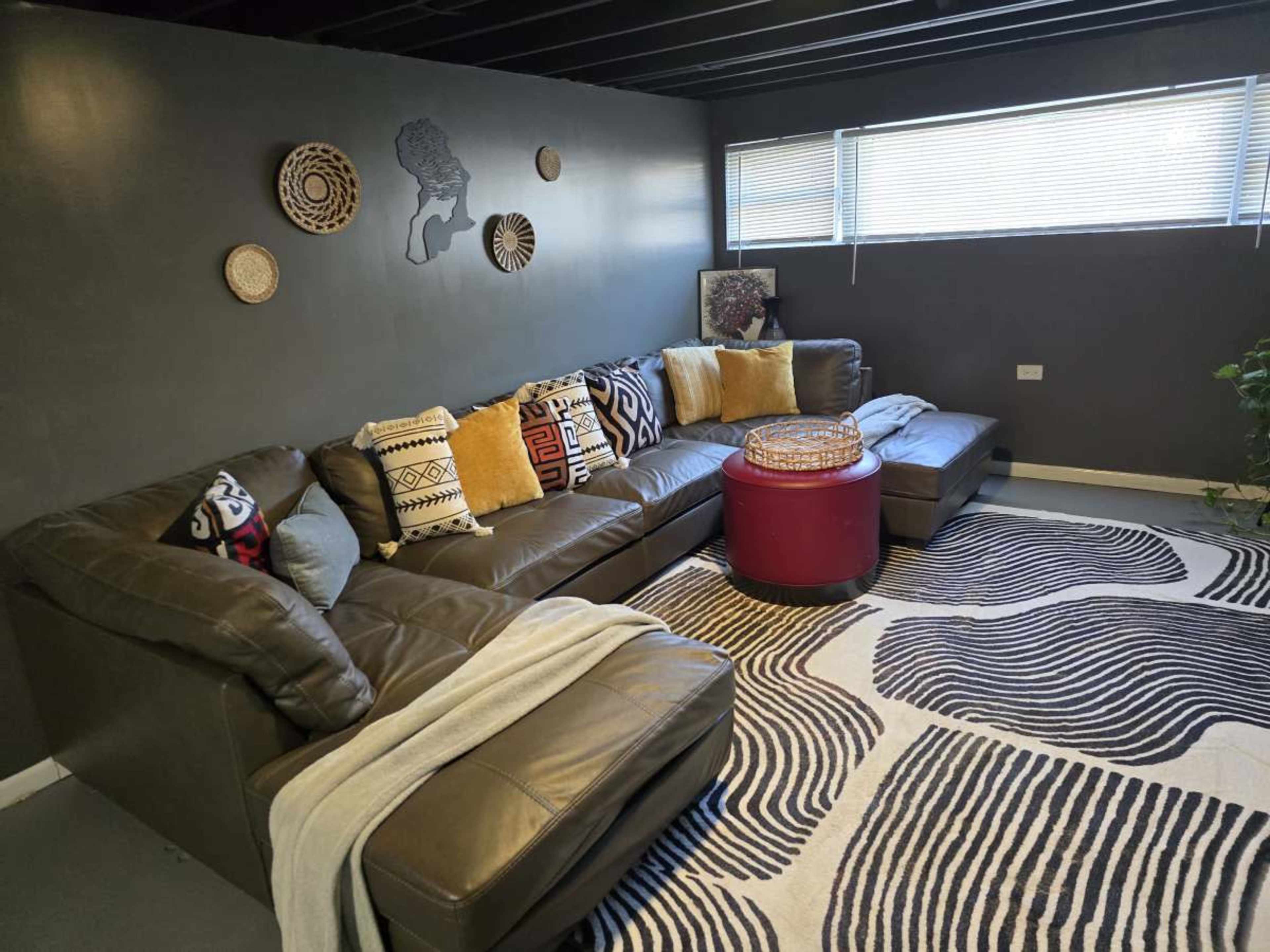 The image shows a modern living room with a sectional leather sofa, decorative cushions, a round red ottoman, and a patterned rug on the floor.