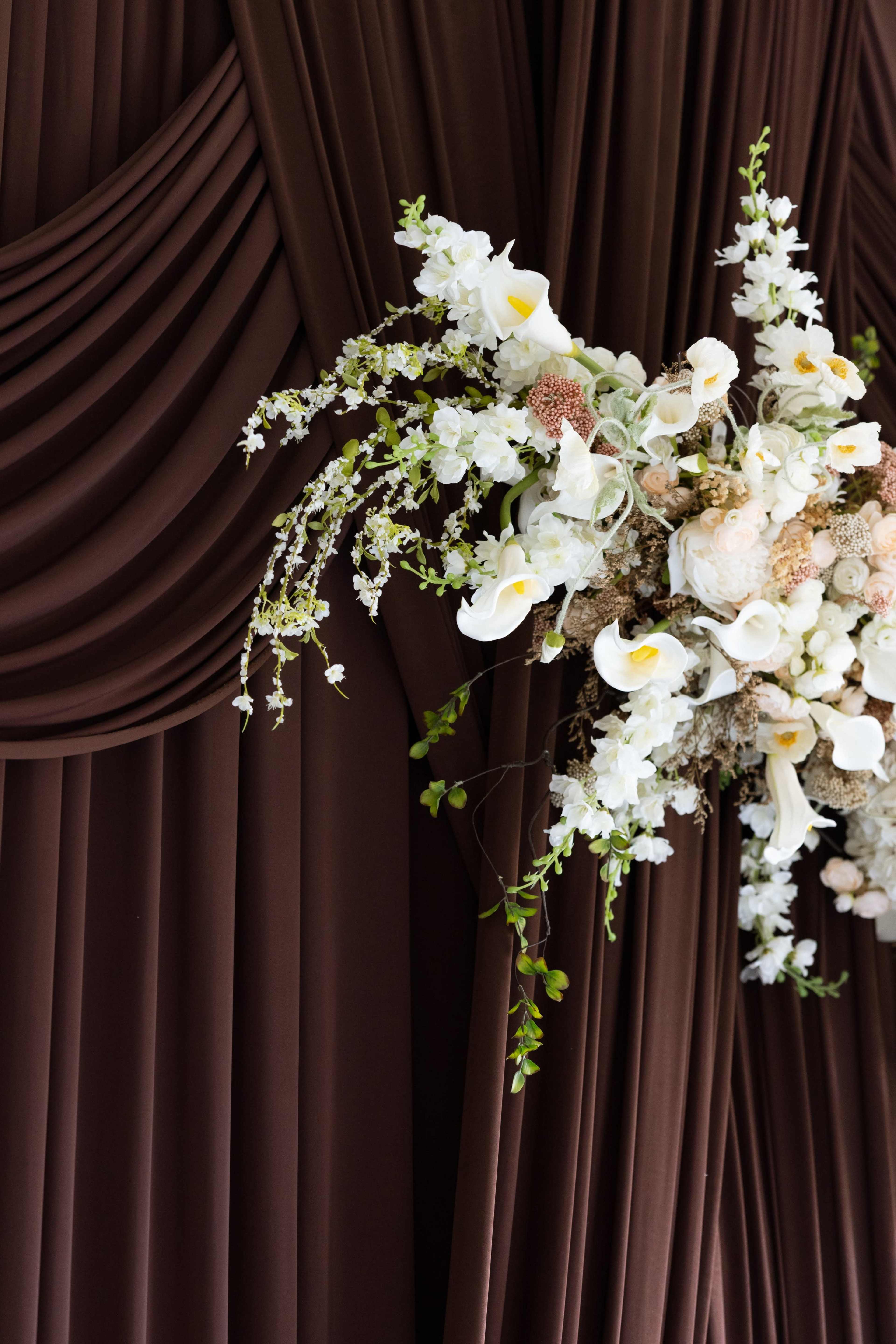 A floral arrangement featuring white and cream flowers is displayed against a backdrop of dark brown draped fabric.
