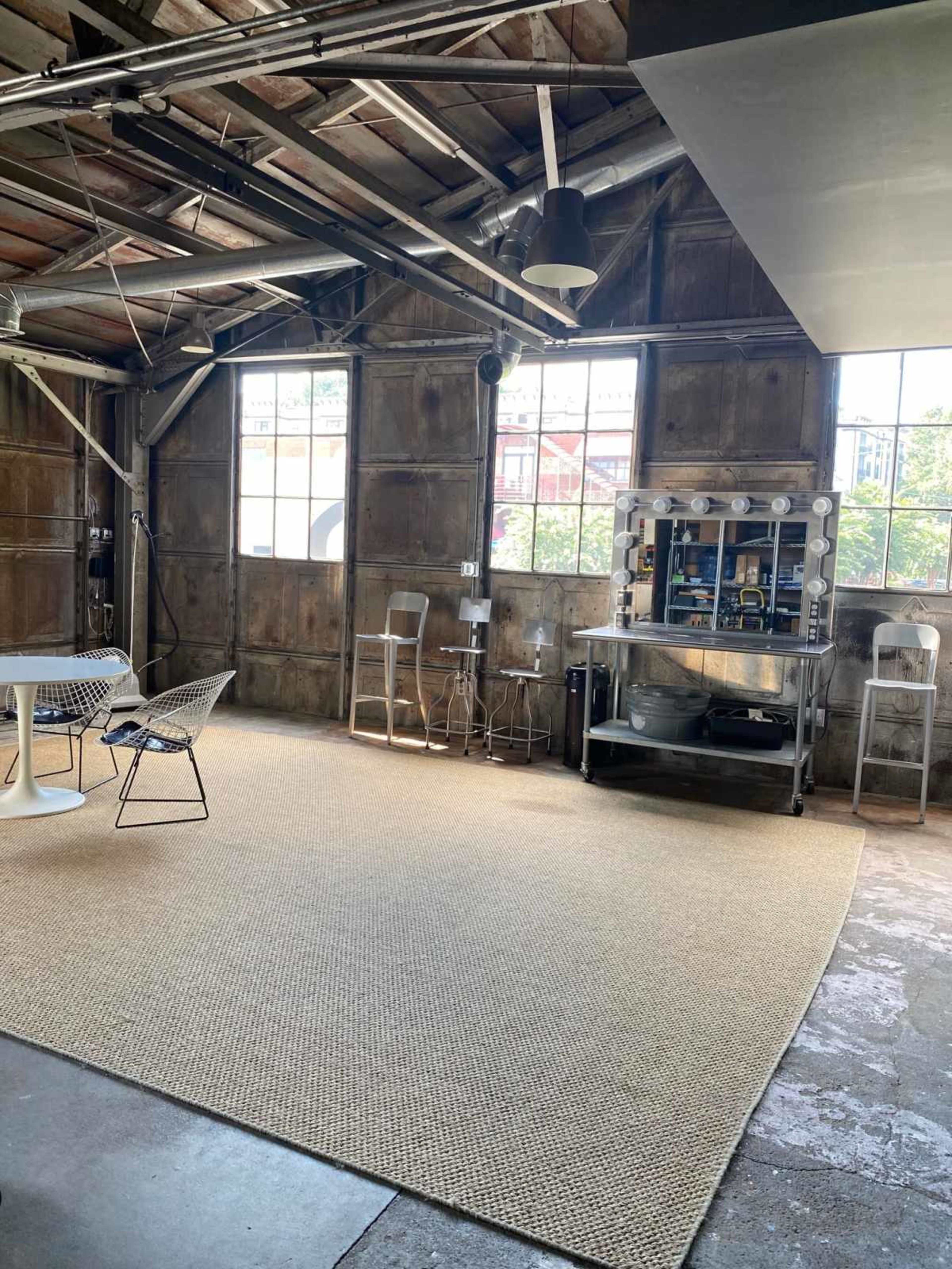 The image shows a spacious industrial-style room with large windows, a beige rug on the floor, and a seating area featuring a round table and metal chairs.