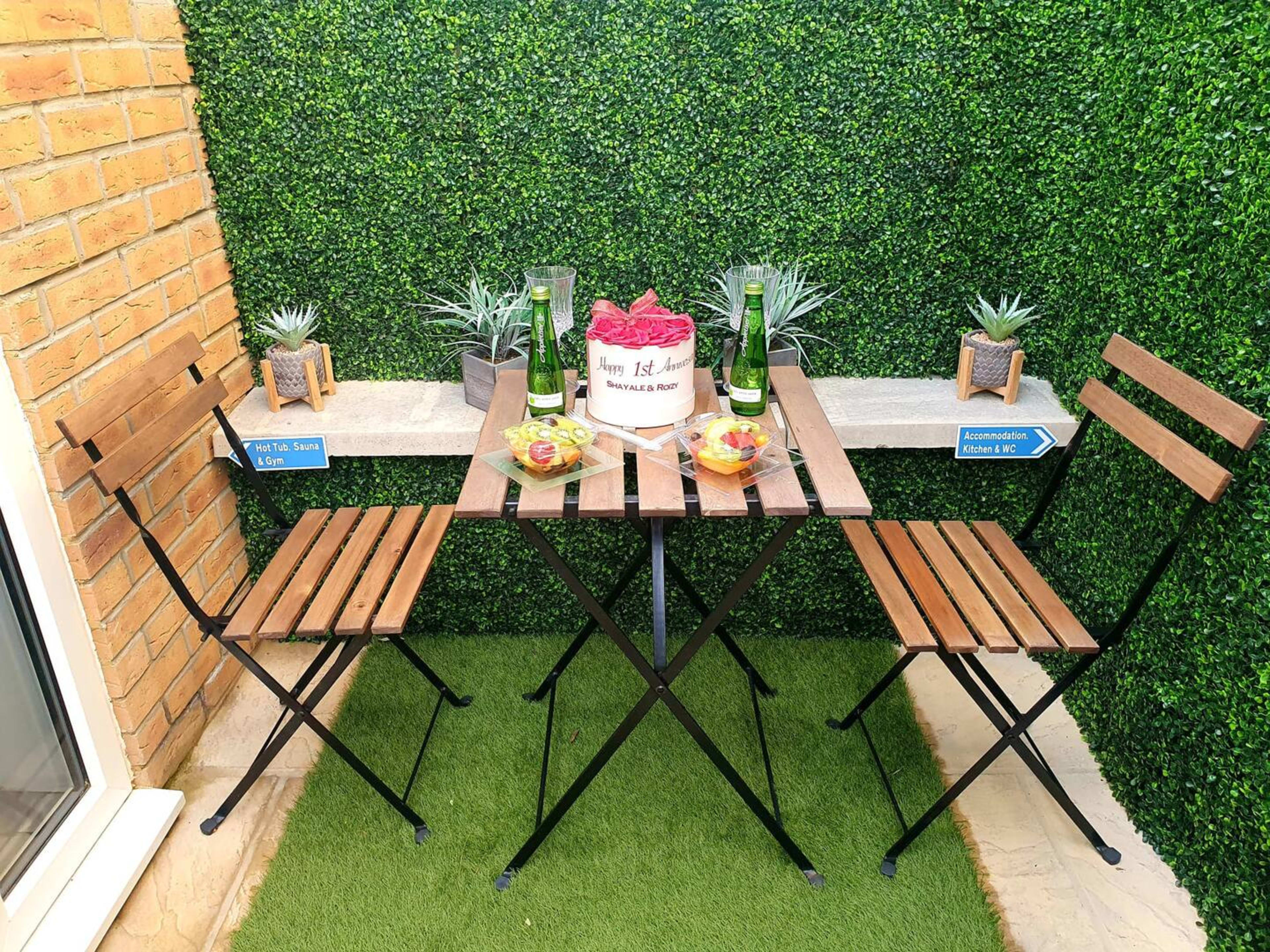 A two-seat table set with a cake and champagne bottles is positioned against a green hedge backdrop.
