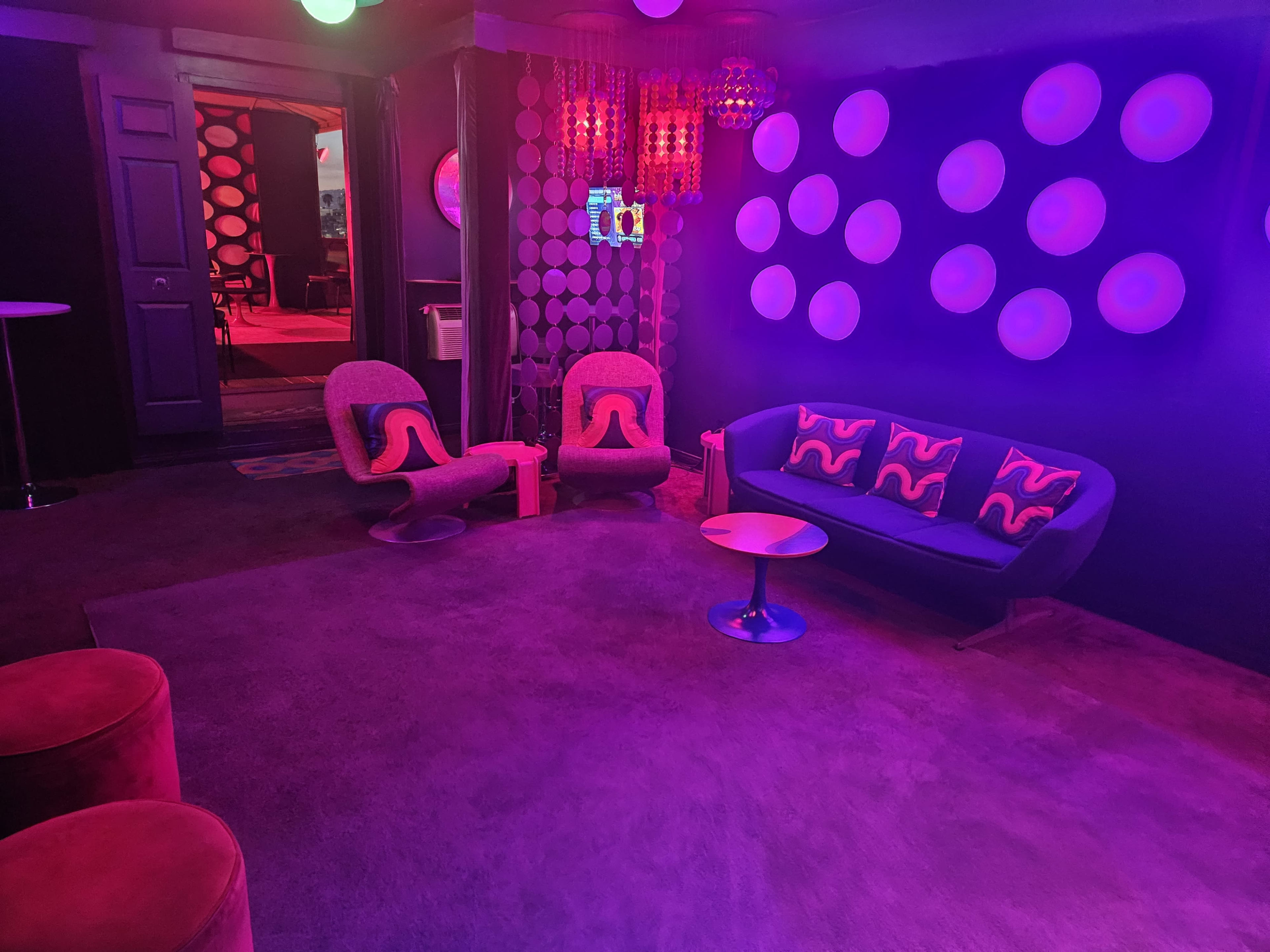 The image shows a modern lounge area with a purple and pink color scheme, featuring a sofa, chairs, a coffee table, and circular wall decorations.