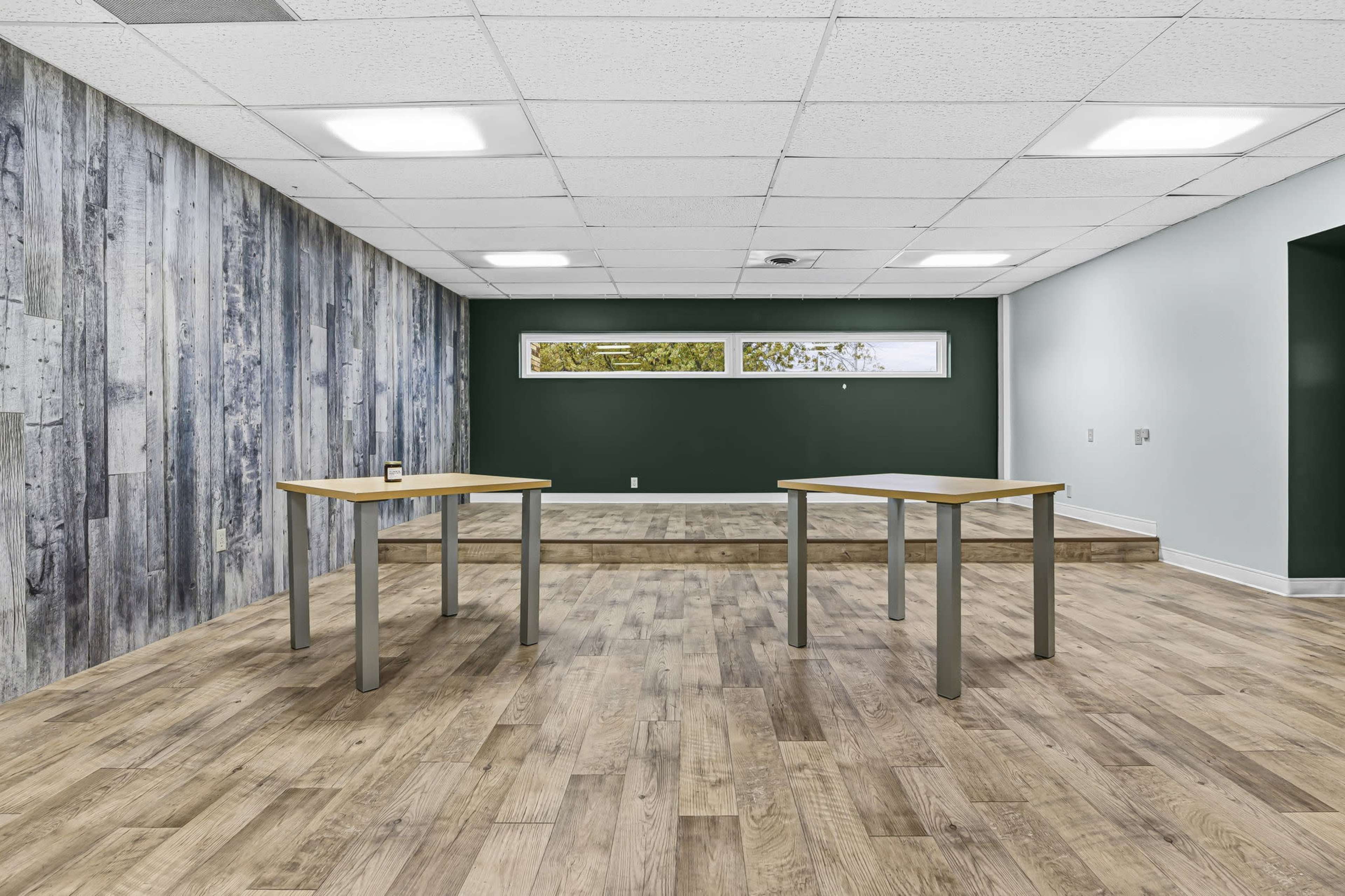 The image depicts a spacious room with two tables, wooden flooring, a green accent wall, and a section raised by stairs at the far end.
