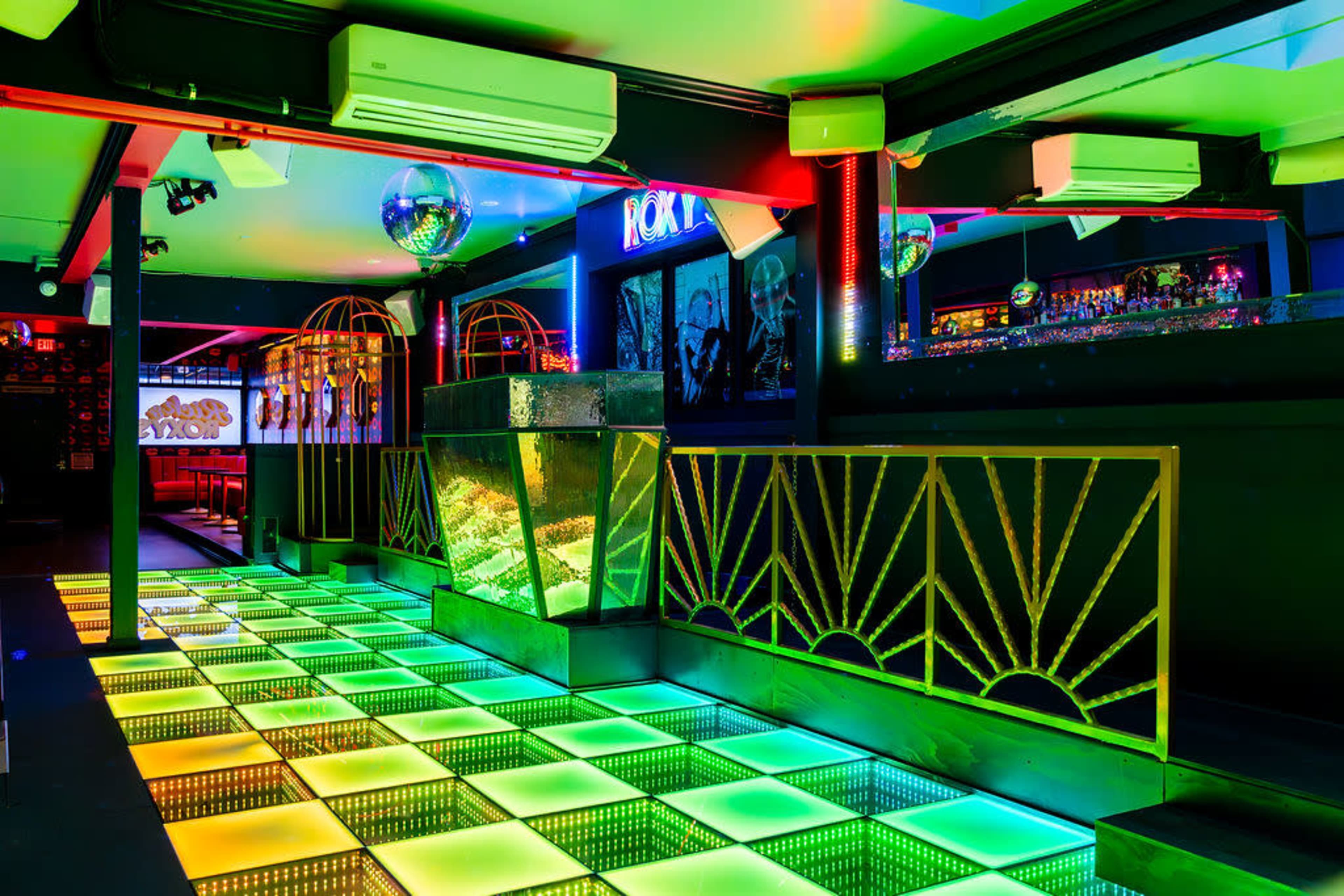 The image shows a brightly lit nightclub interior featuring a colorful illuminated dance floor with a mirrored disco ball and stylish decor.