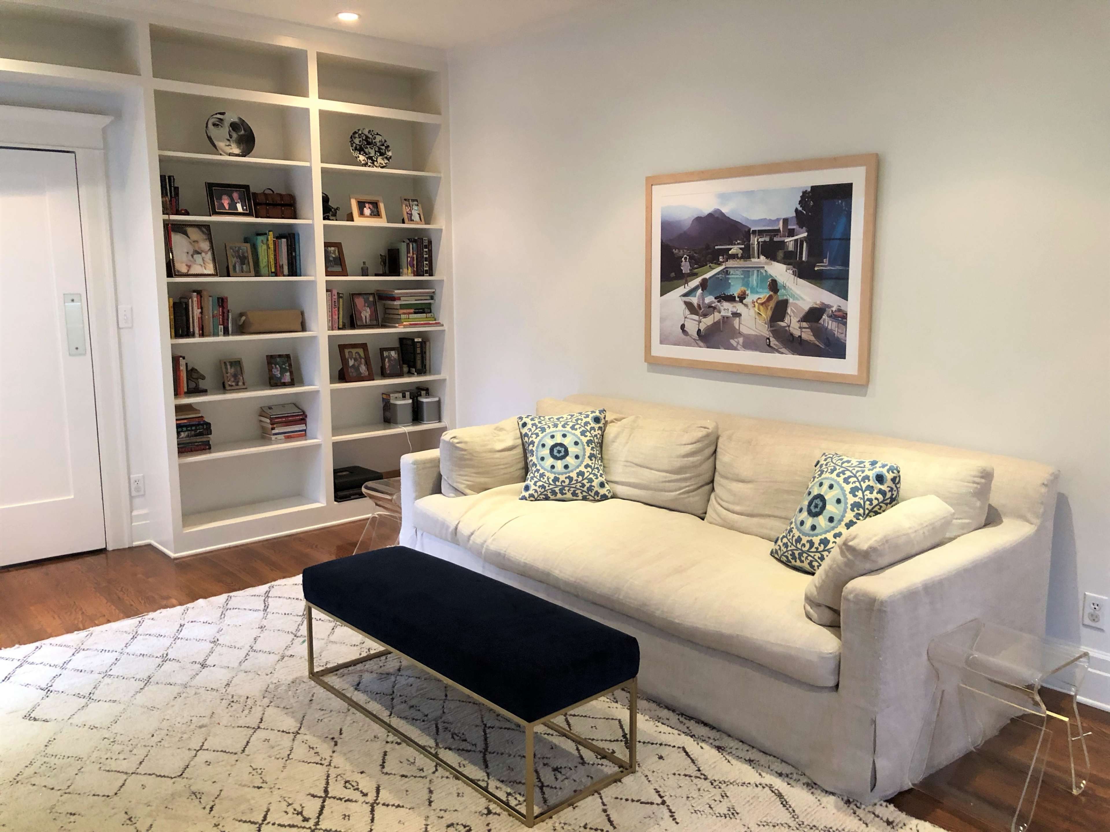 A light-colored living room features a sofa with decorative cushions, a dark bench, and a bookshelf filled with various books and objects.