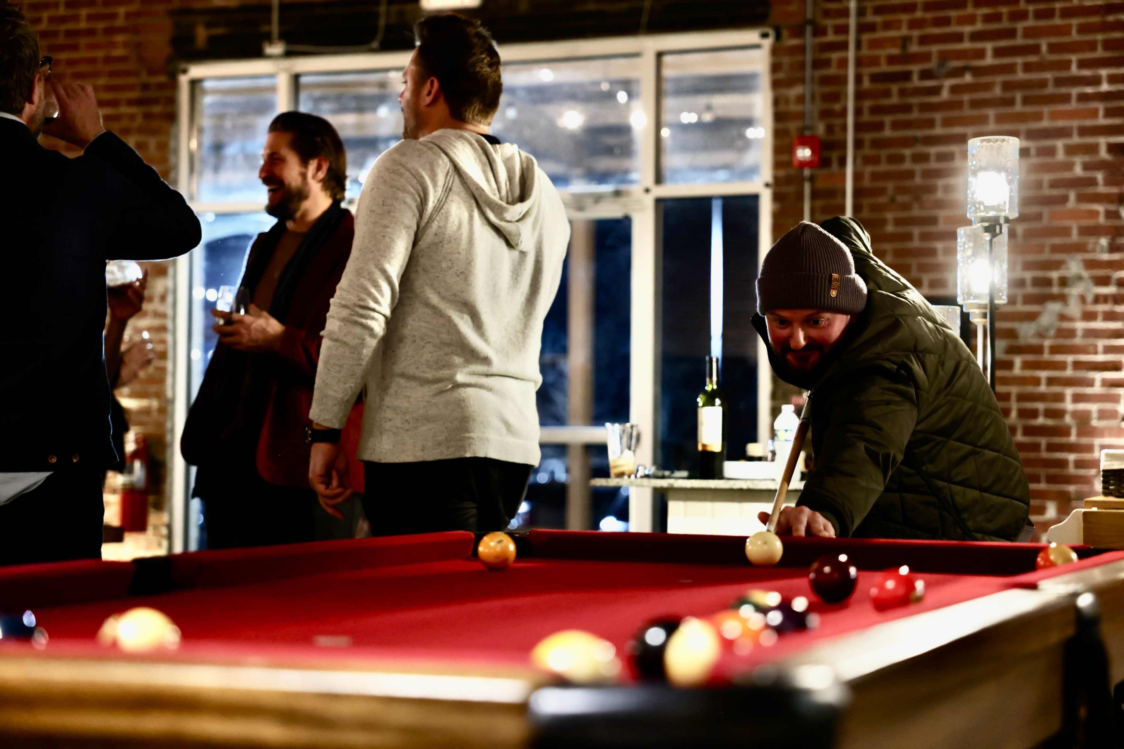 A group of people socializes near a pool table, while one player prepares to take a shot.