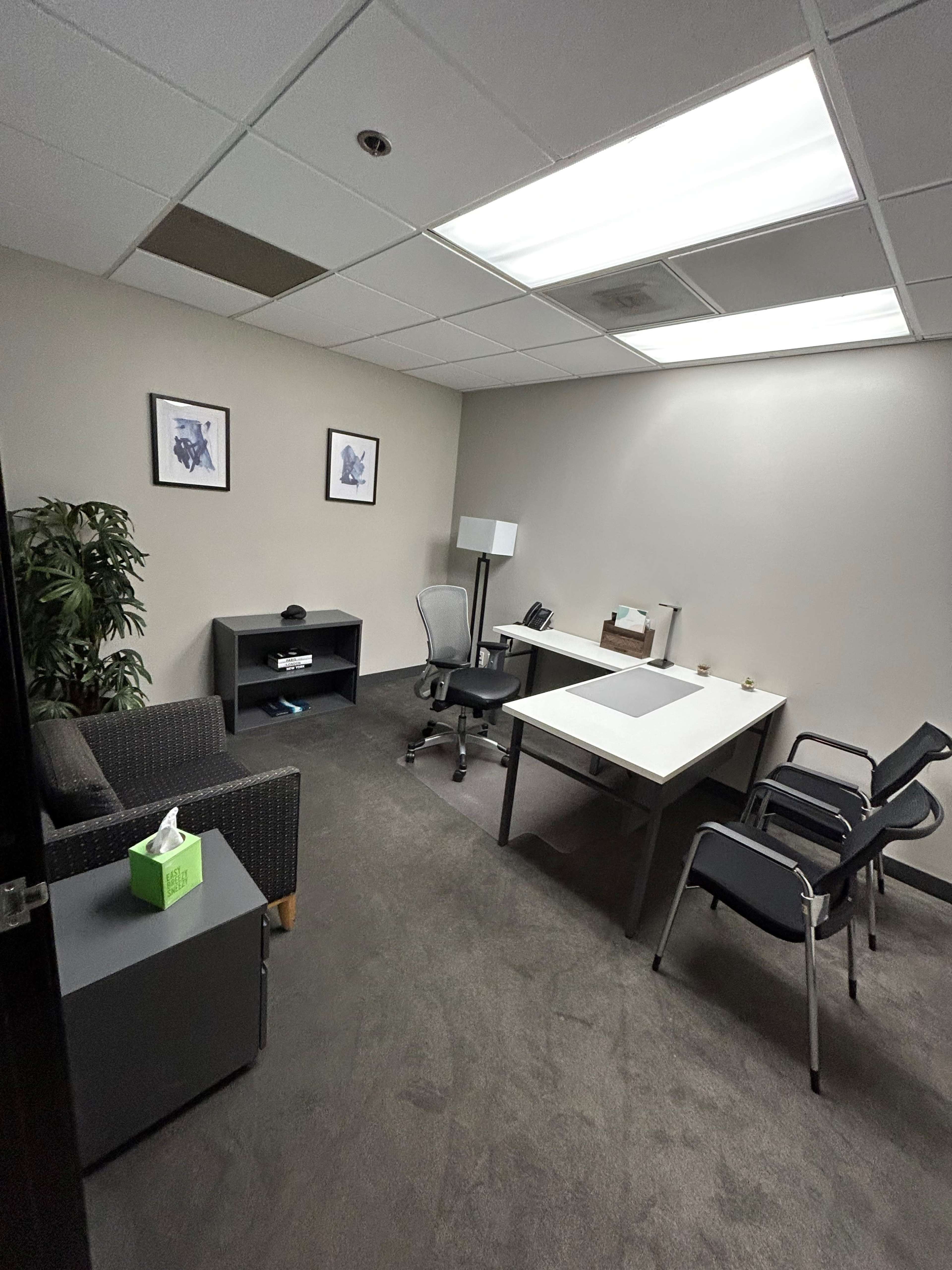 The image shows a small office with a desk, a chair, a small couch, and a few framed pictures on the wall.