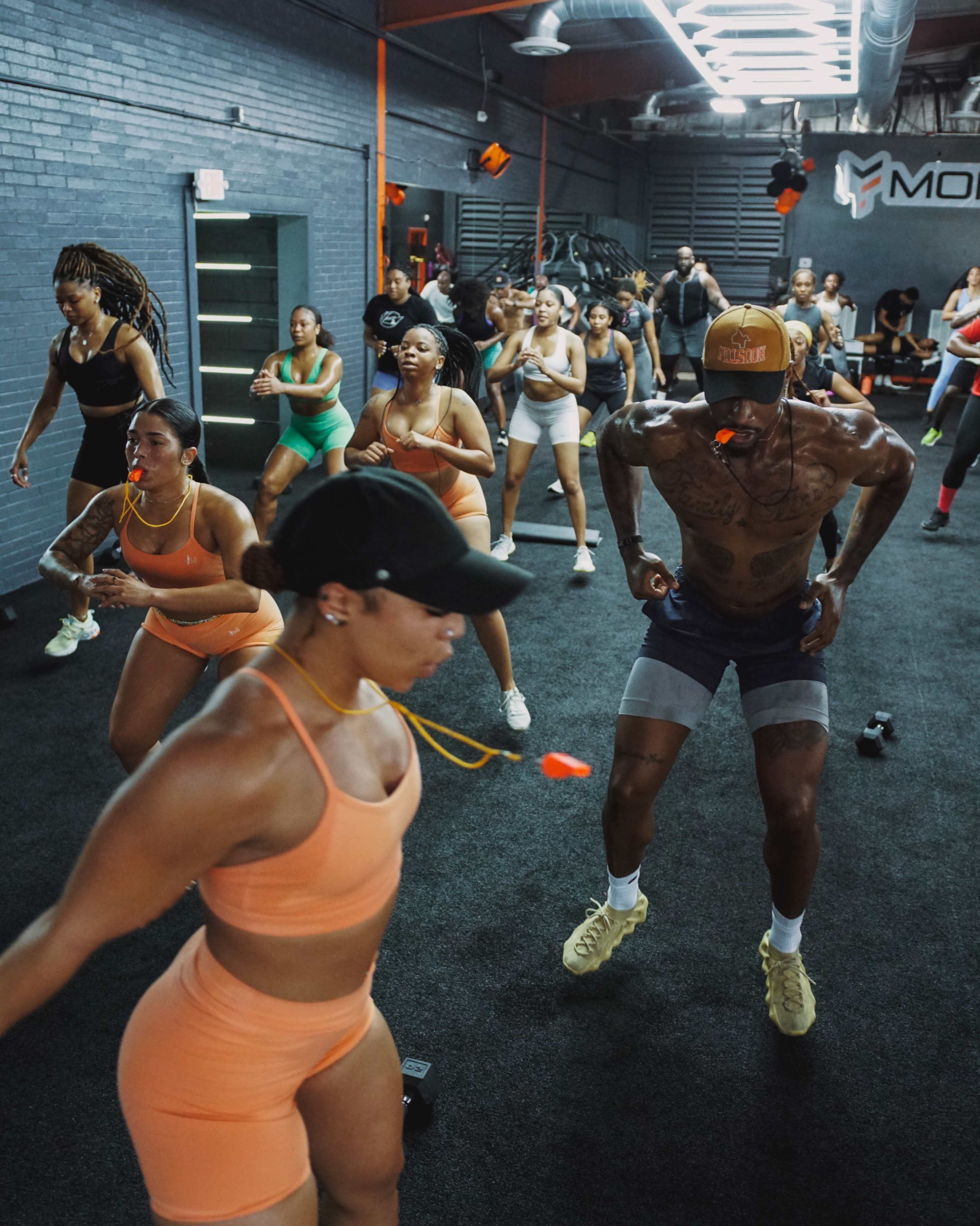 A group of people are engaged in a high-intensity workout session in a gym, with some wearing athletic clothing and others focused on their routines.
