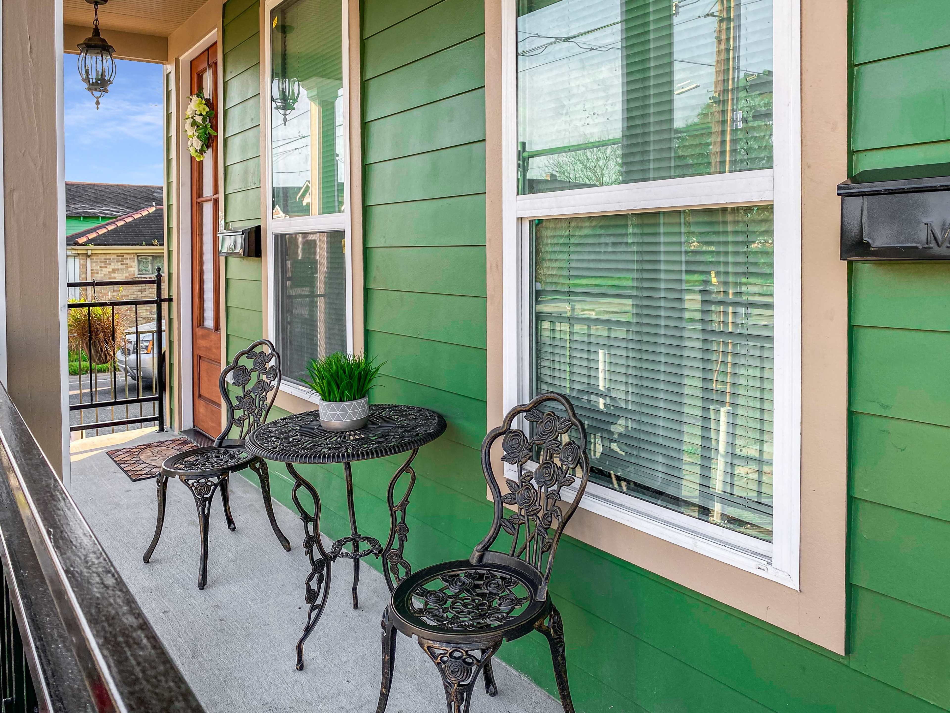 A small porch with a green exterior, featuring a round table and two metal chairs, and decorative plants near the windows.