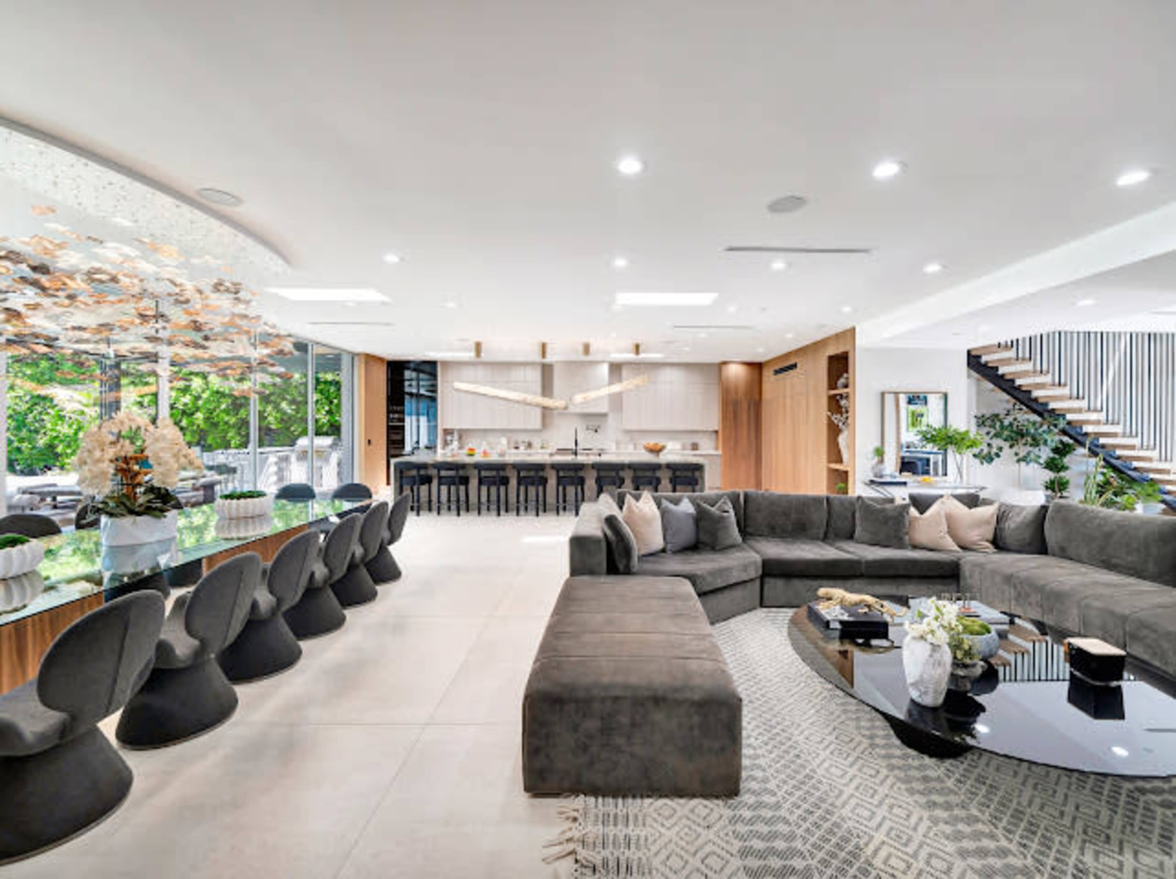 The image shows a modern, open-concept living and dining area featuring a large sectional sofa, a sleek dining table with unique chairs, and a staircase in the background.