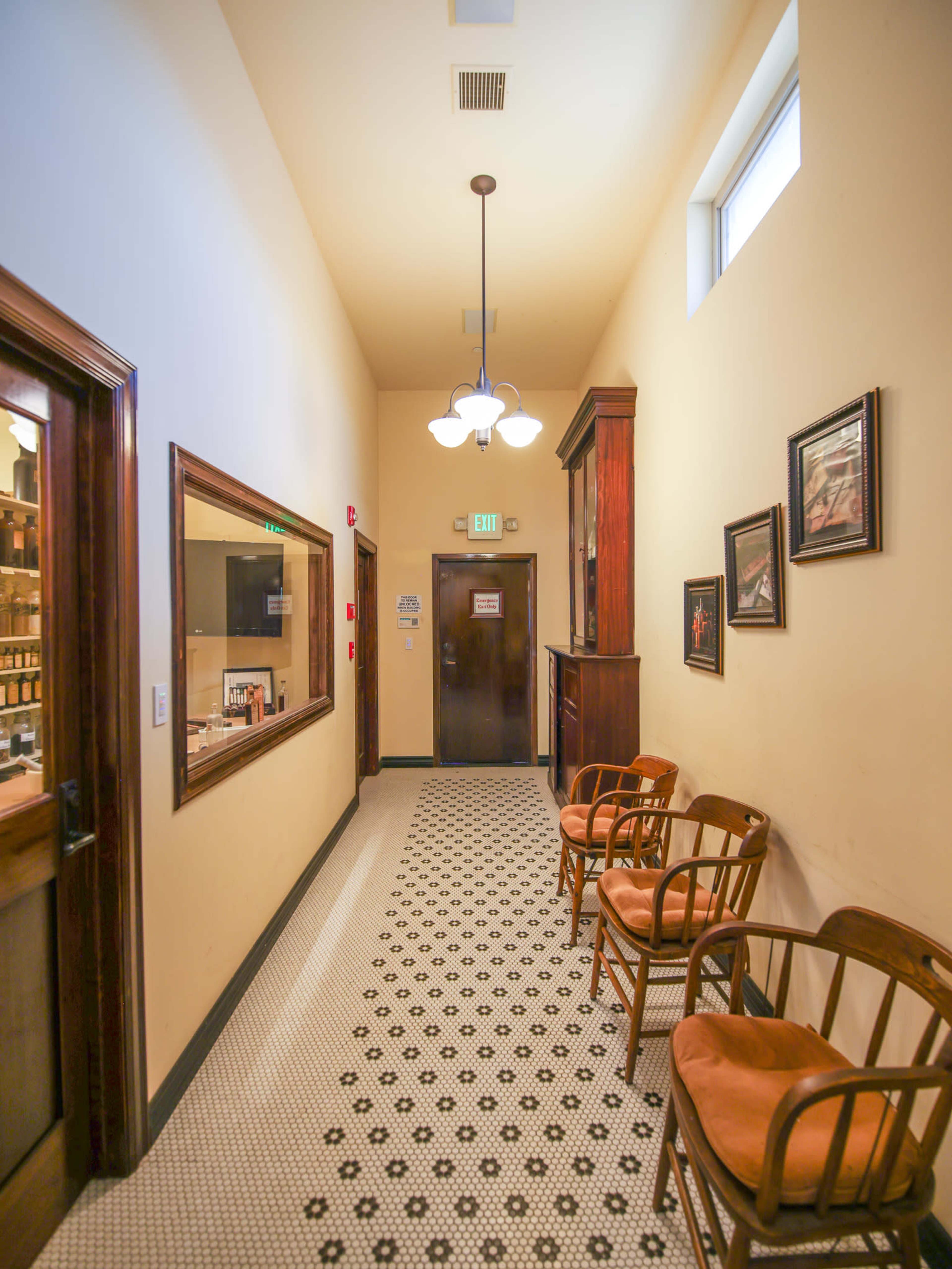 A narrow hallway features wooden chairs along one side, with framed artworks on the walls and a door at the far end.