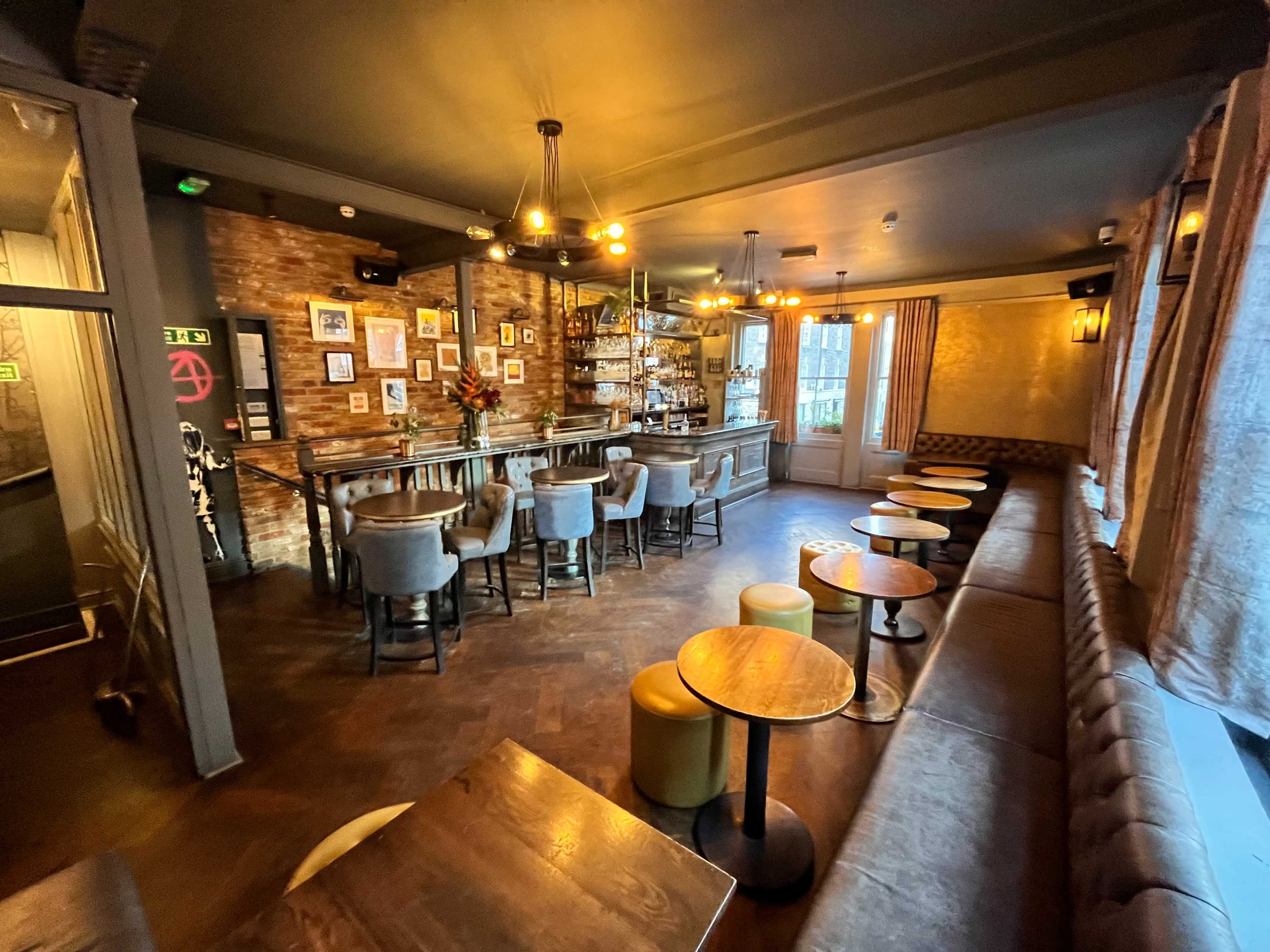 The image shows a dimly lit bar interior with a wooden counter, high stools, and tables arranged along a leather-upholstered bench.
