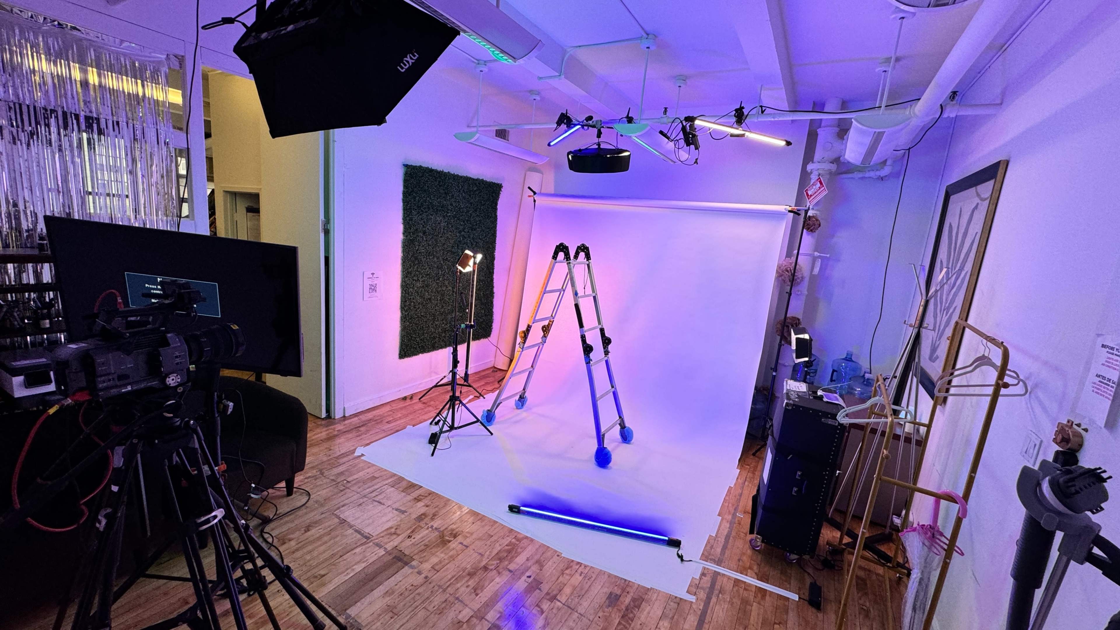 A photography studio setup with a white backdrop, lighting equipment, a ladder, and a camera.