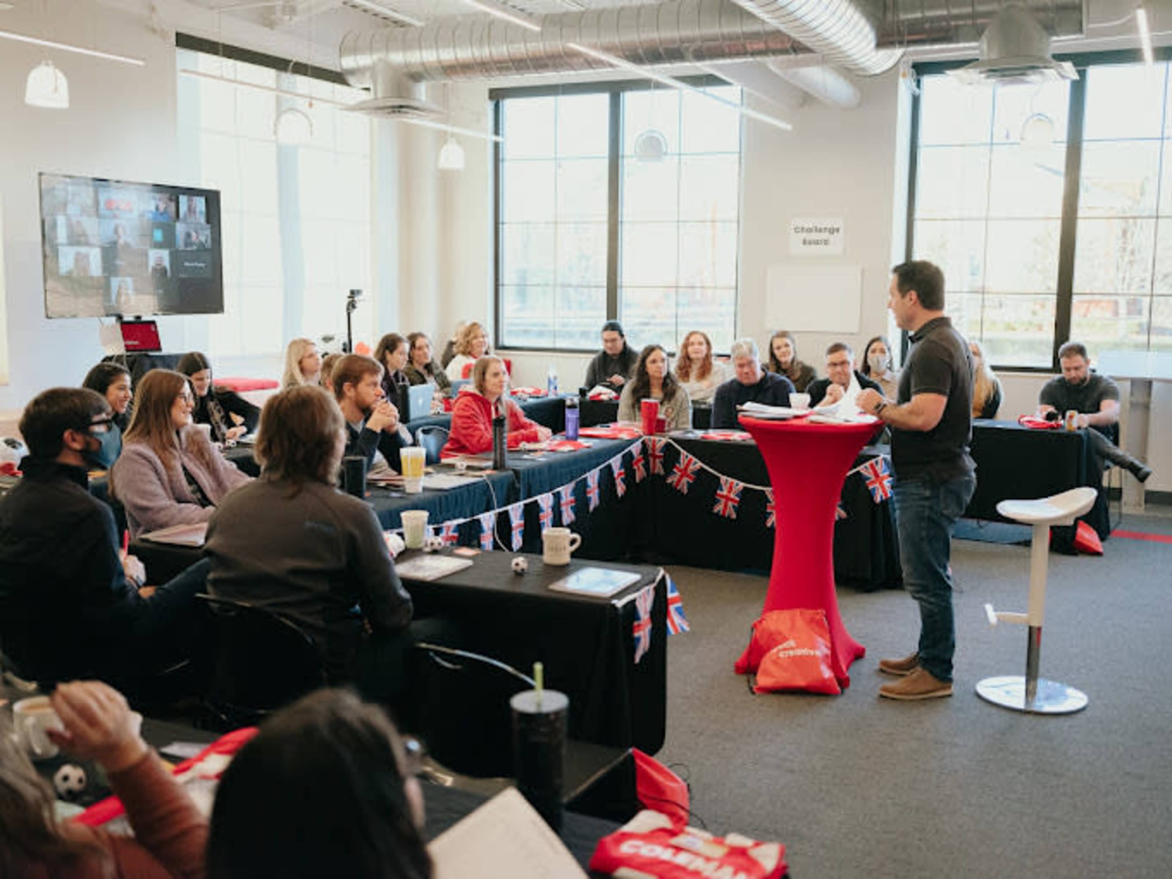 A speaker stands in front of an audience in a classroom setting, with participants seated at tables decorated with British flags.