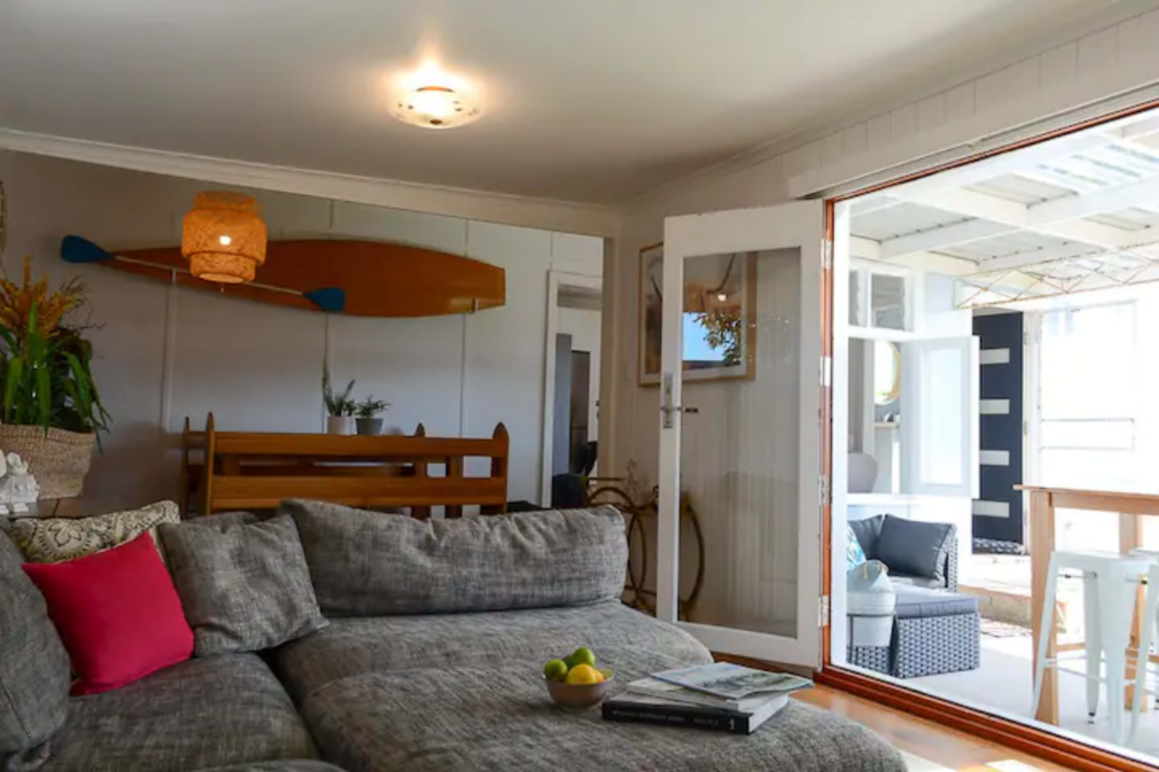 A cozy living area features a gray sectional sofa, a decorative paddle on the wall, and doors opening to a bright outdoor patio.