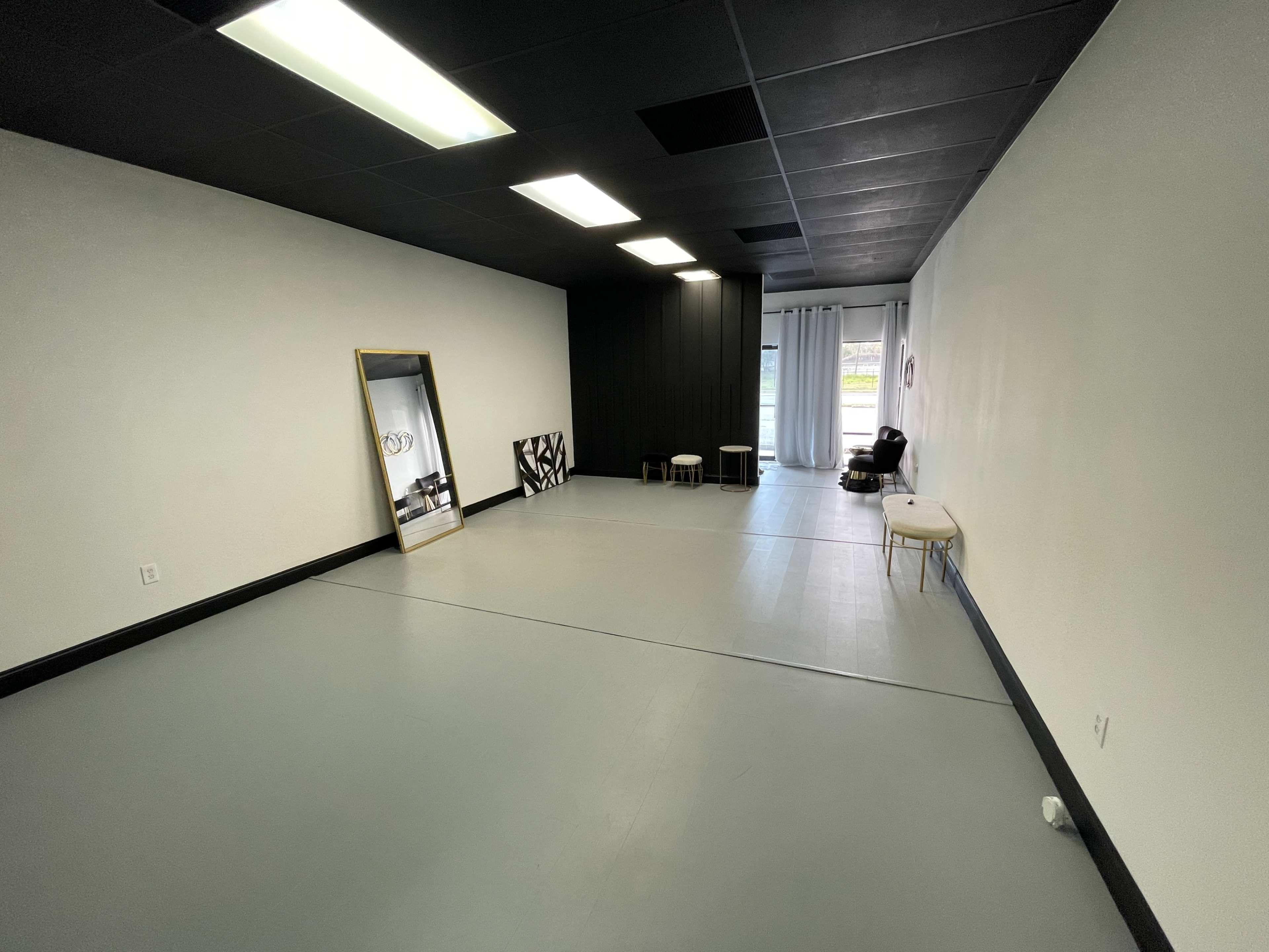 An empty, well-lit room with gray flooring, a large mirror, two small stools, and a curtain window.