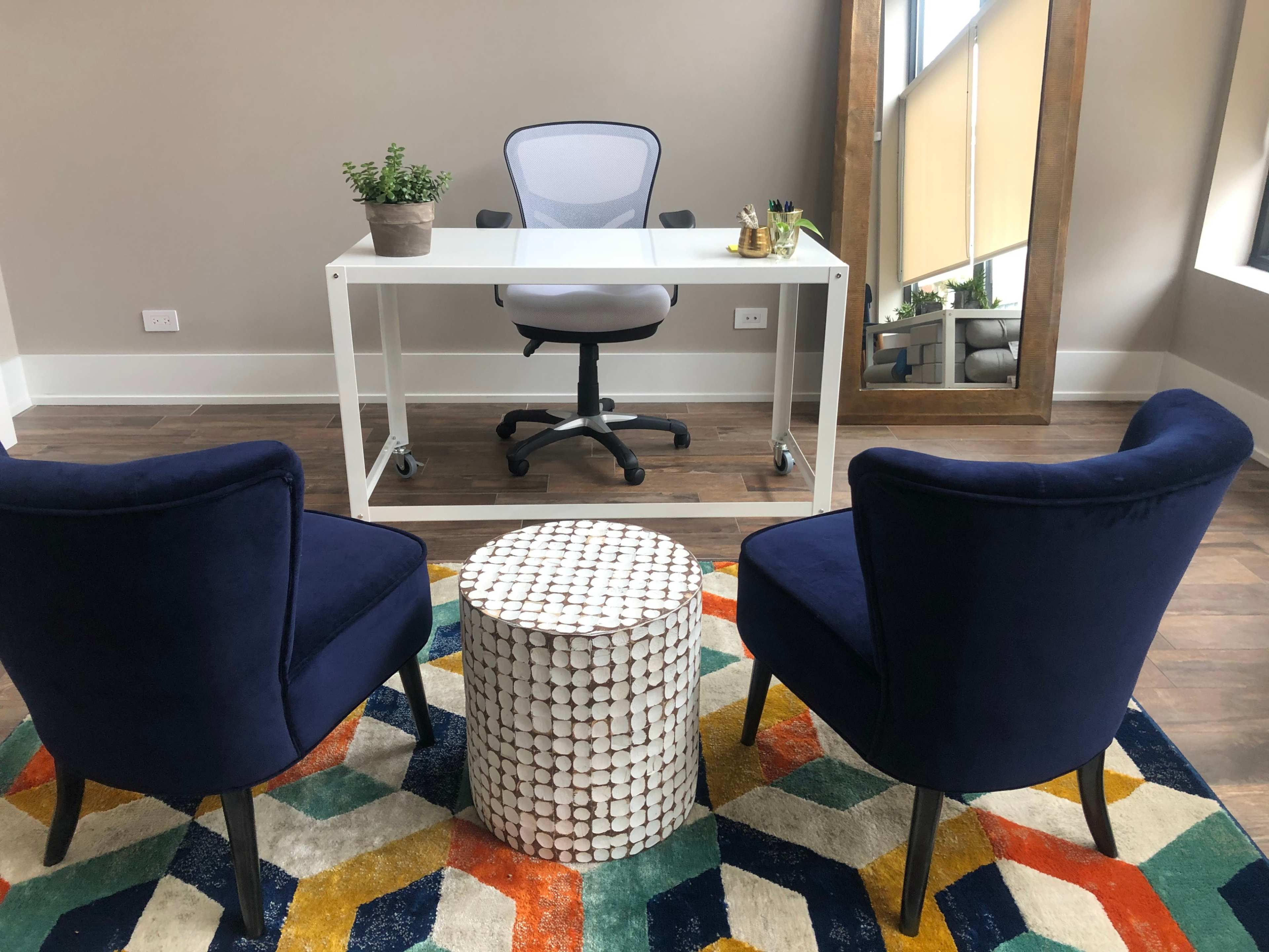 A modern office space featuring a white desk with a chair, two blue upholstered chairs, and a round, patterned table on a colorful rug.