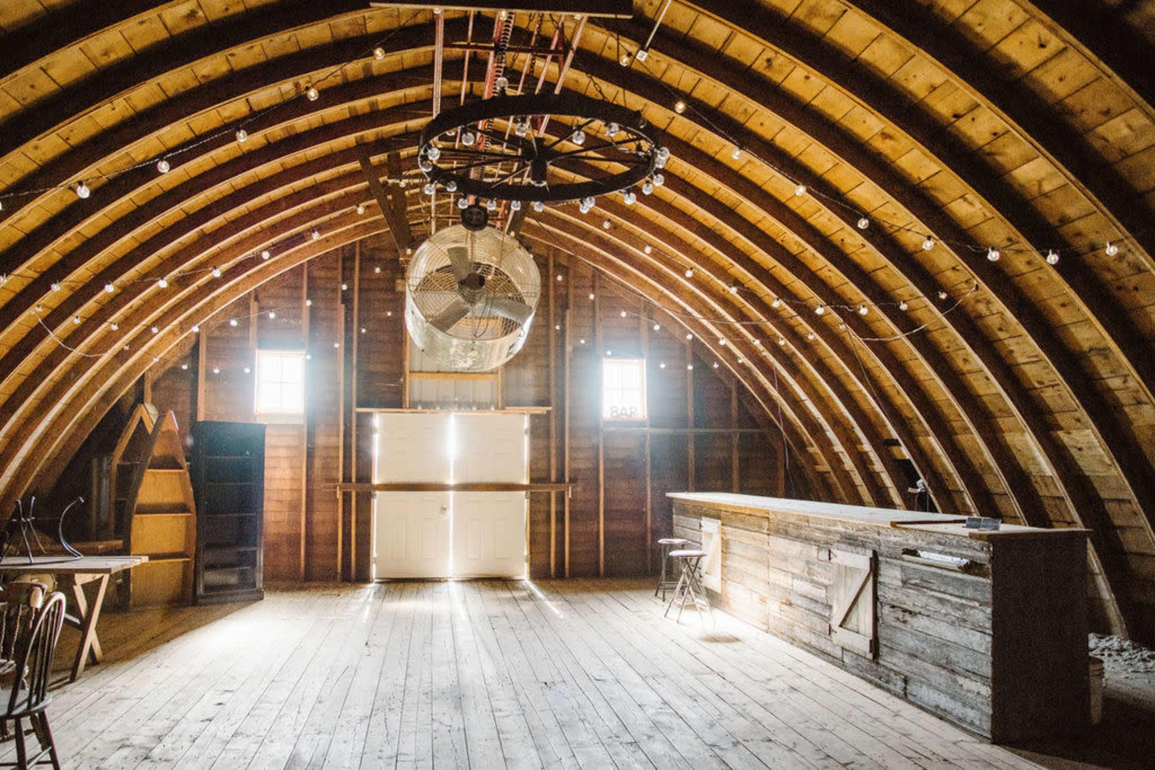 The interior of a rustic barn features arched wooden beams, large windows, a ceiling fan, and a wooden bar area.