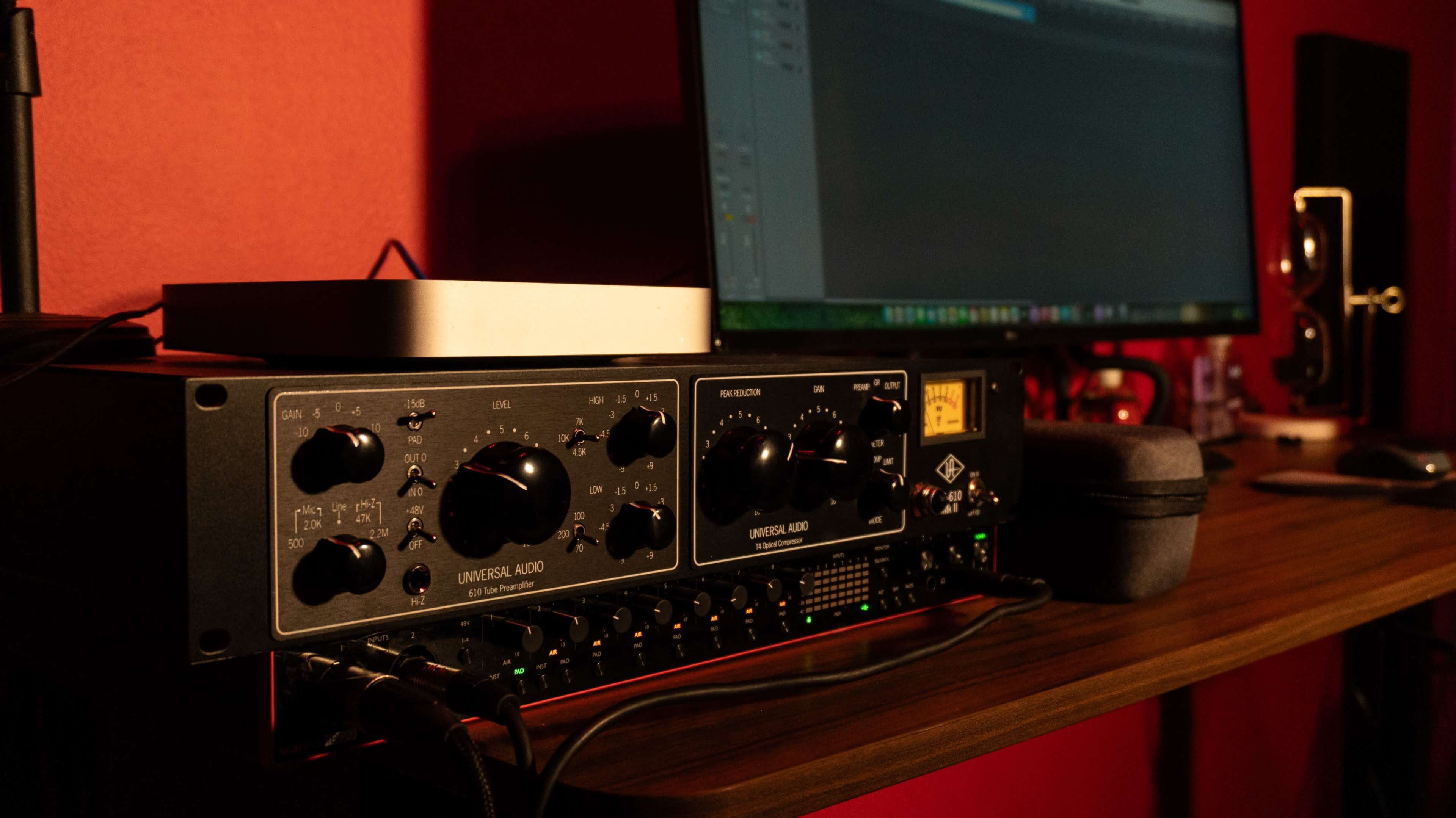 A Universal Audio audio interface is placed on a wooden desk next to a computer monitor displaying audio editing software.