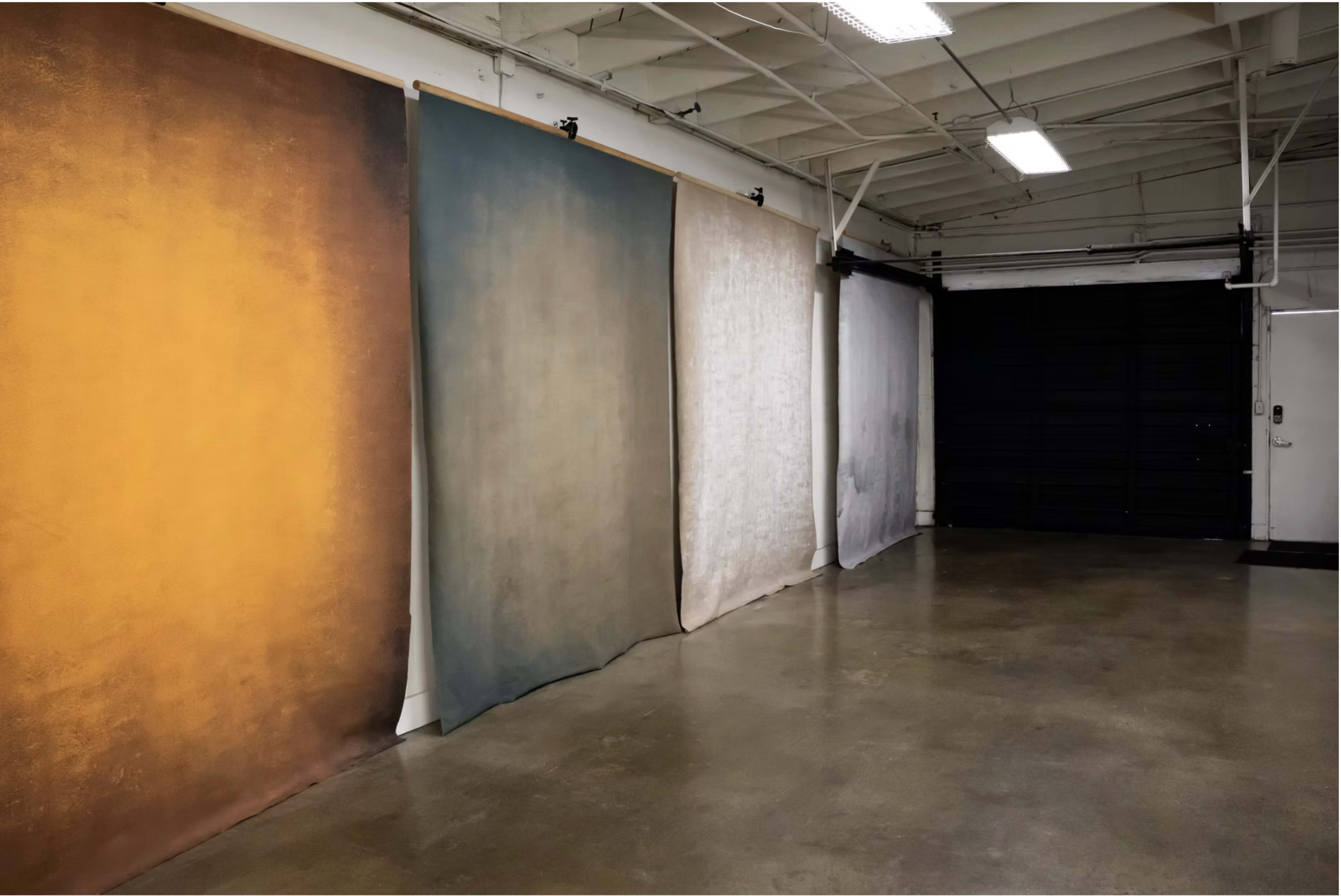 The image shows an art studio with four large fabric backdrops in varying colors, positioned against a concrete floor and white walls.