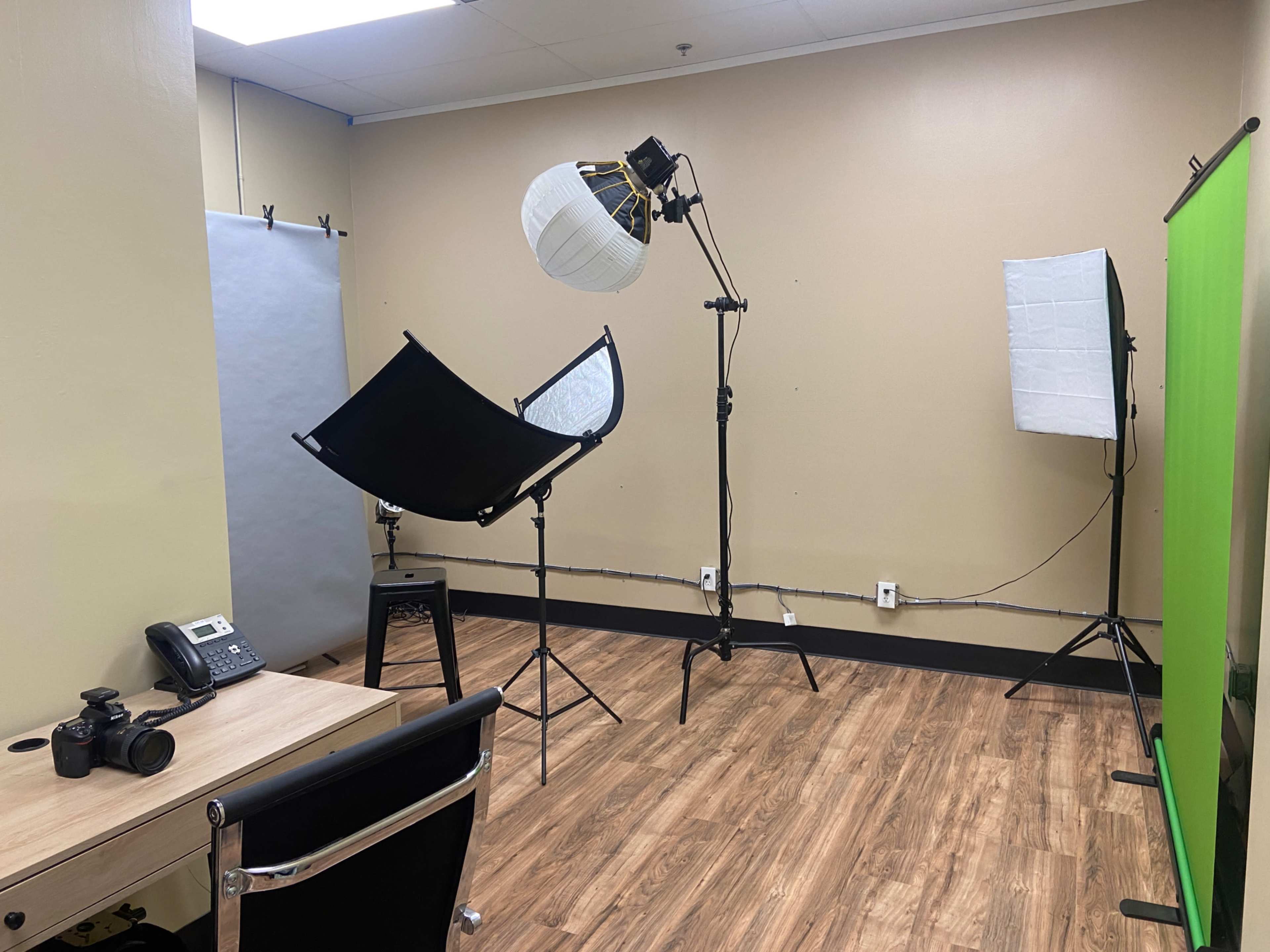 A small studio space equipped with photography lights, a green screen, a camera, a phone, and a table.