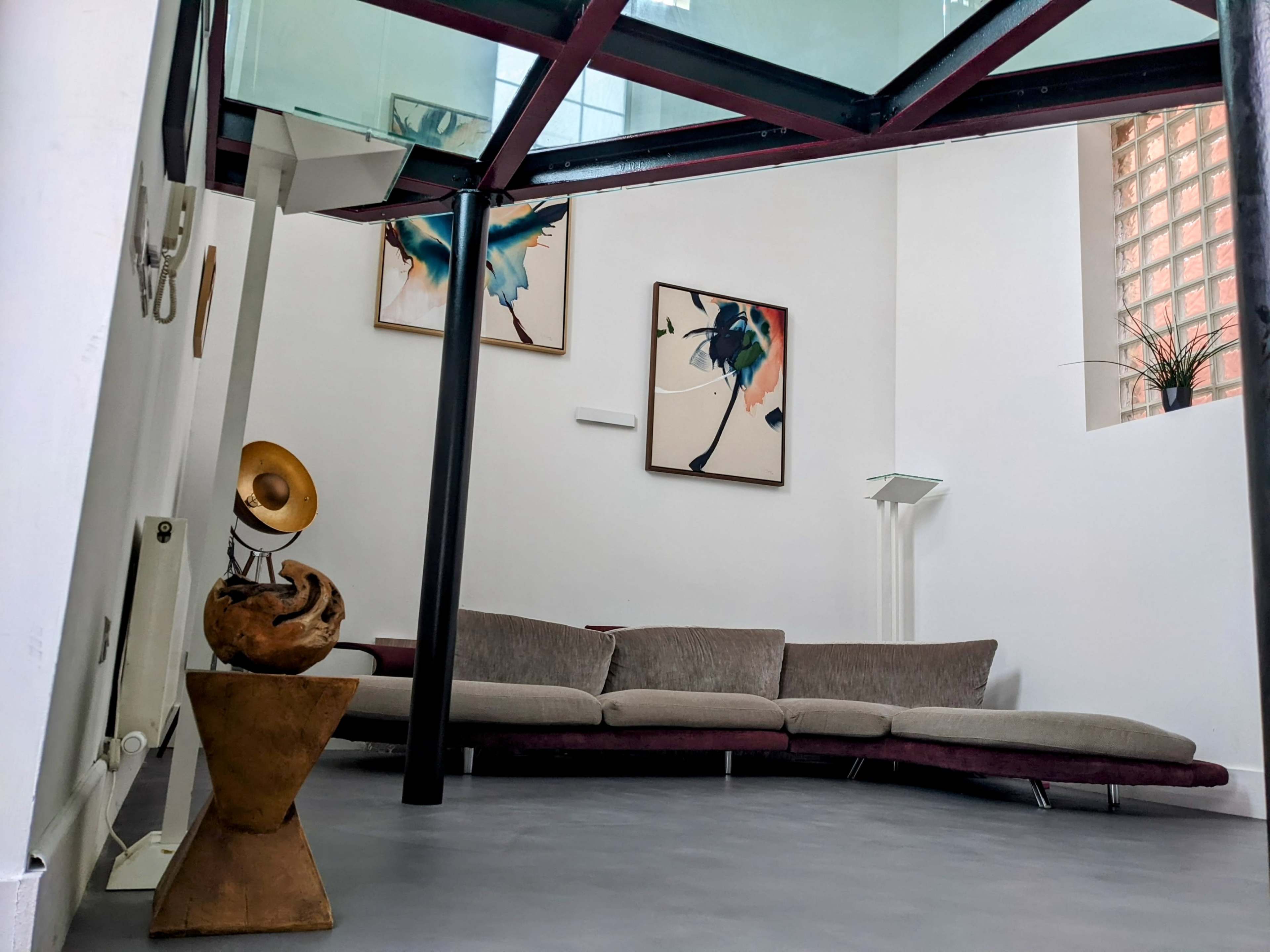 A modern interior with a curved gray sofa, two abstract paintings on the wall, and a glass floor above supported by black pillars.