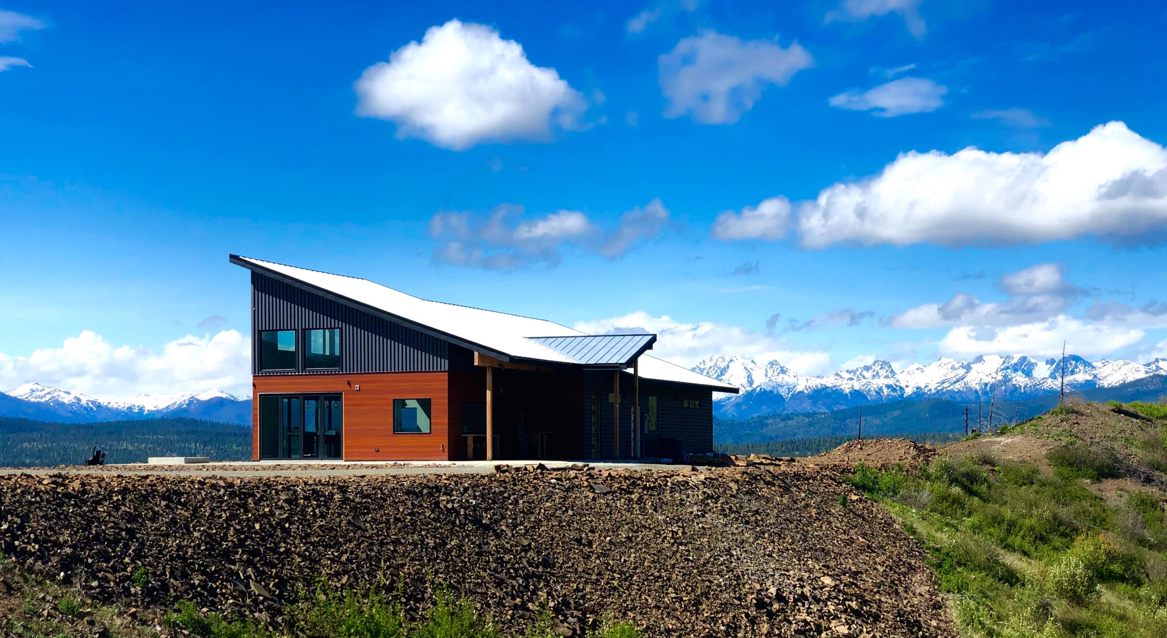 A modern house with a sloped roof situated on a rocky hillside, surrounded by snow-capped mountains under a blue sky with scattered clouds.