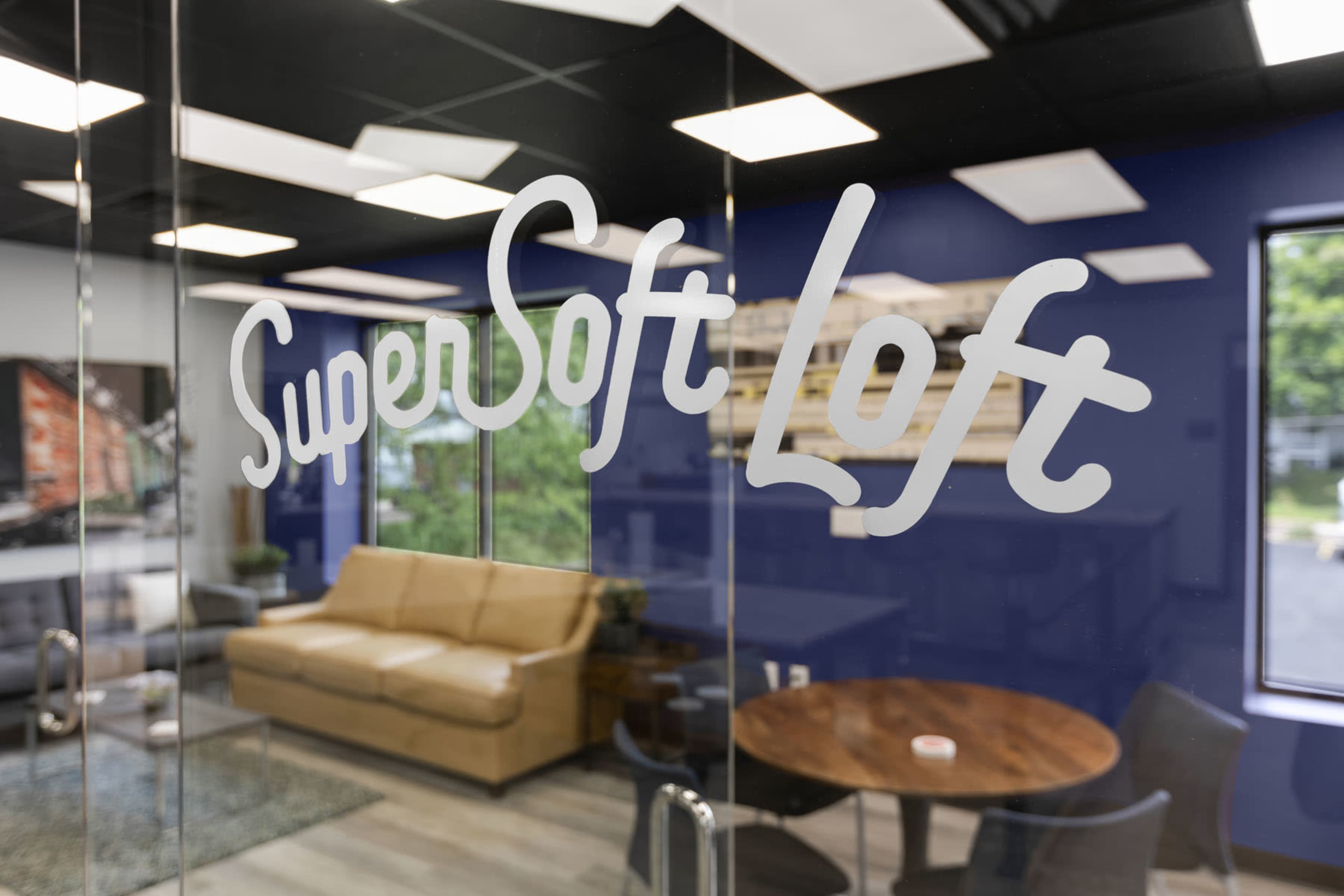 A glass door with the words "SuperSoft Loft" written on it, leading into a modern office space featuring a sofa and a round table.