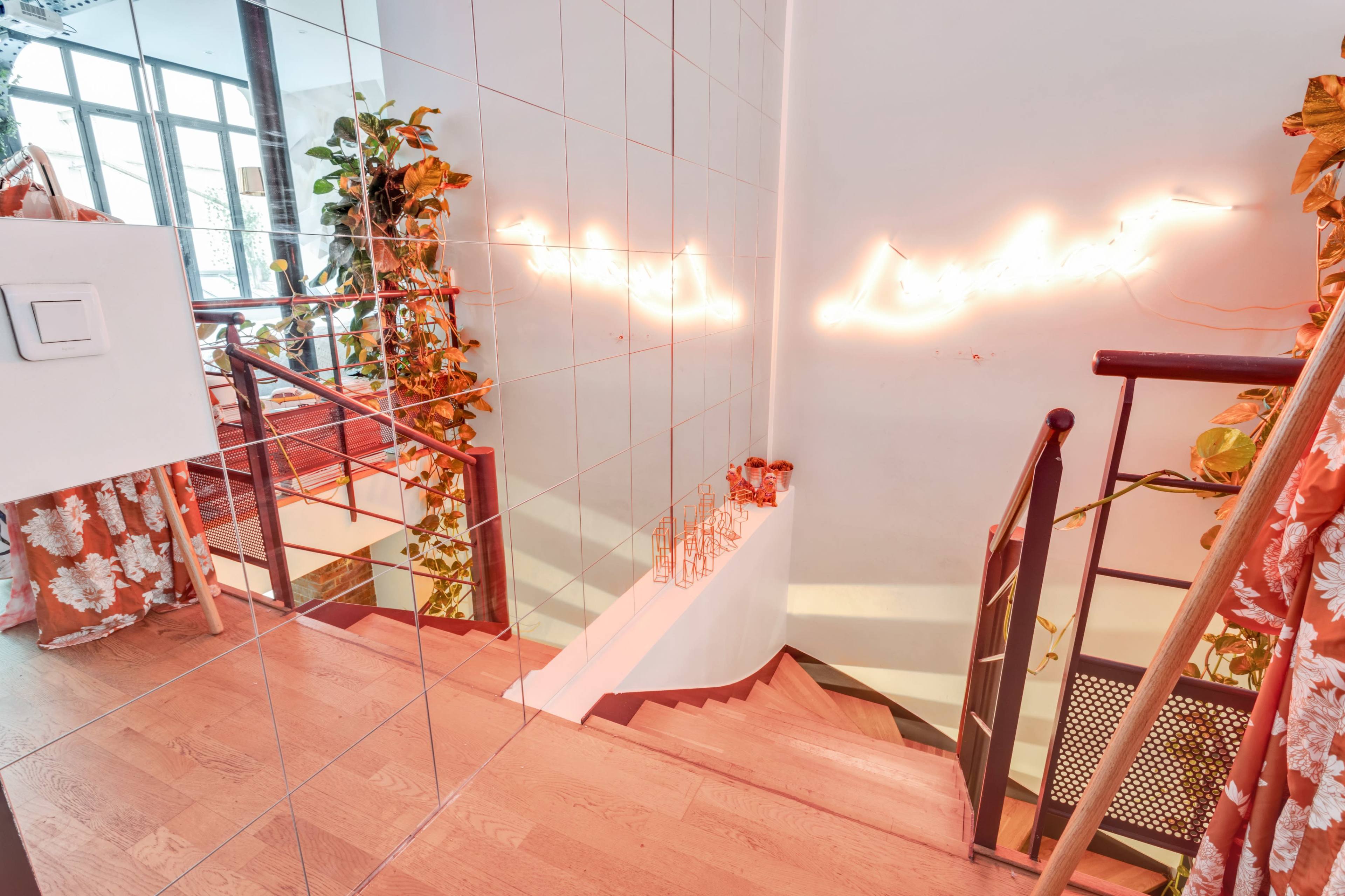 A modern staircase with wooden steps, surrounded by mirrored walls and plants, features a neon sign on the wall above.