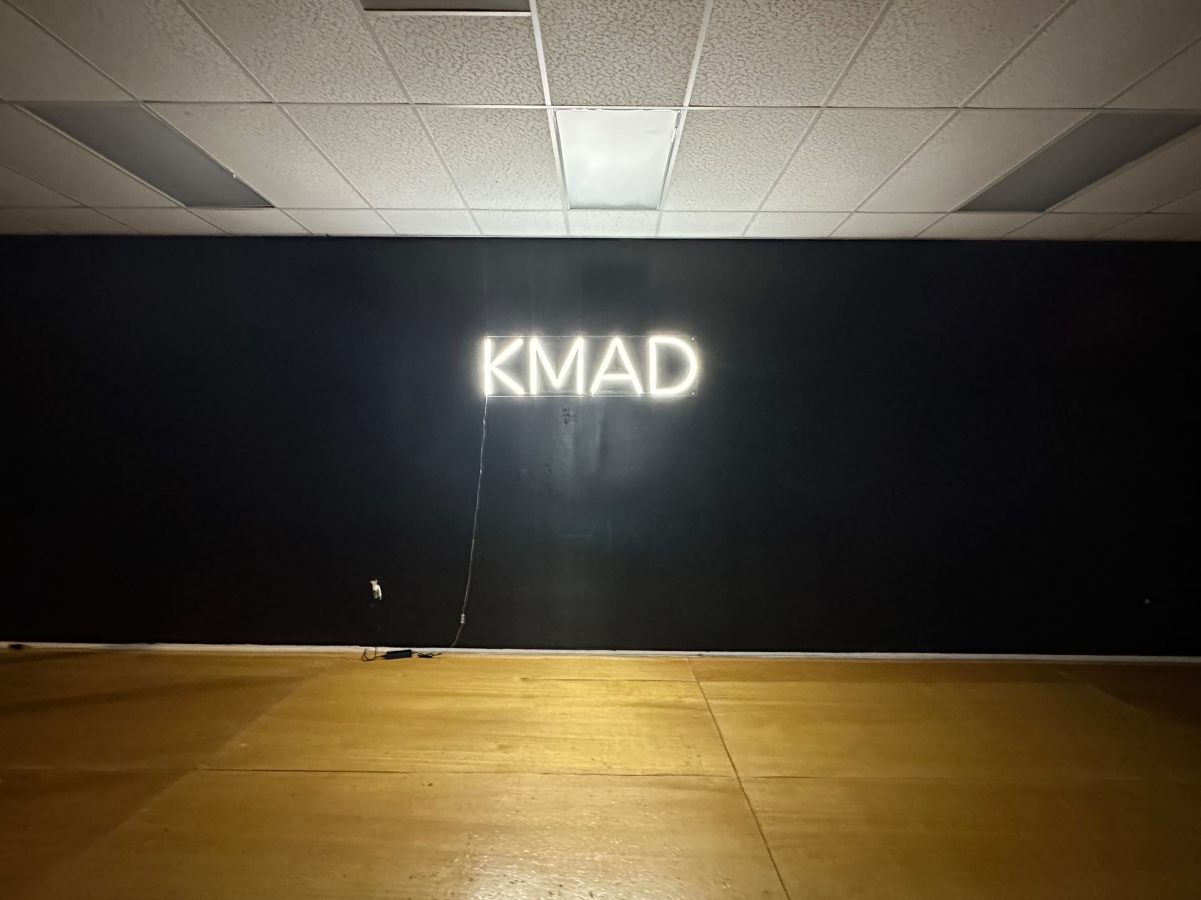 A neon sign displaying the letters "KMAD" is mounted on a dark wall in a sparsely furnished room with wooden flooring.