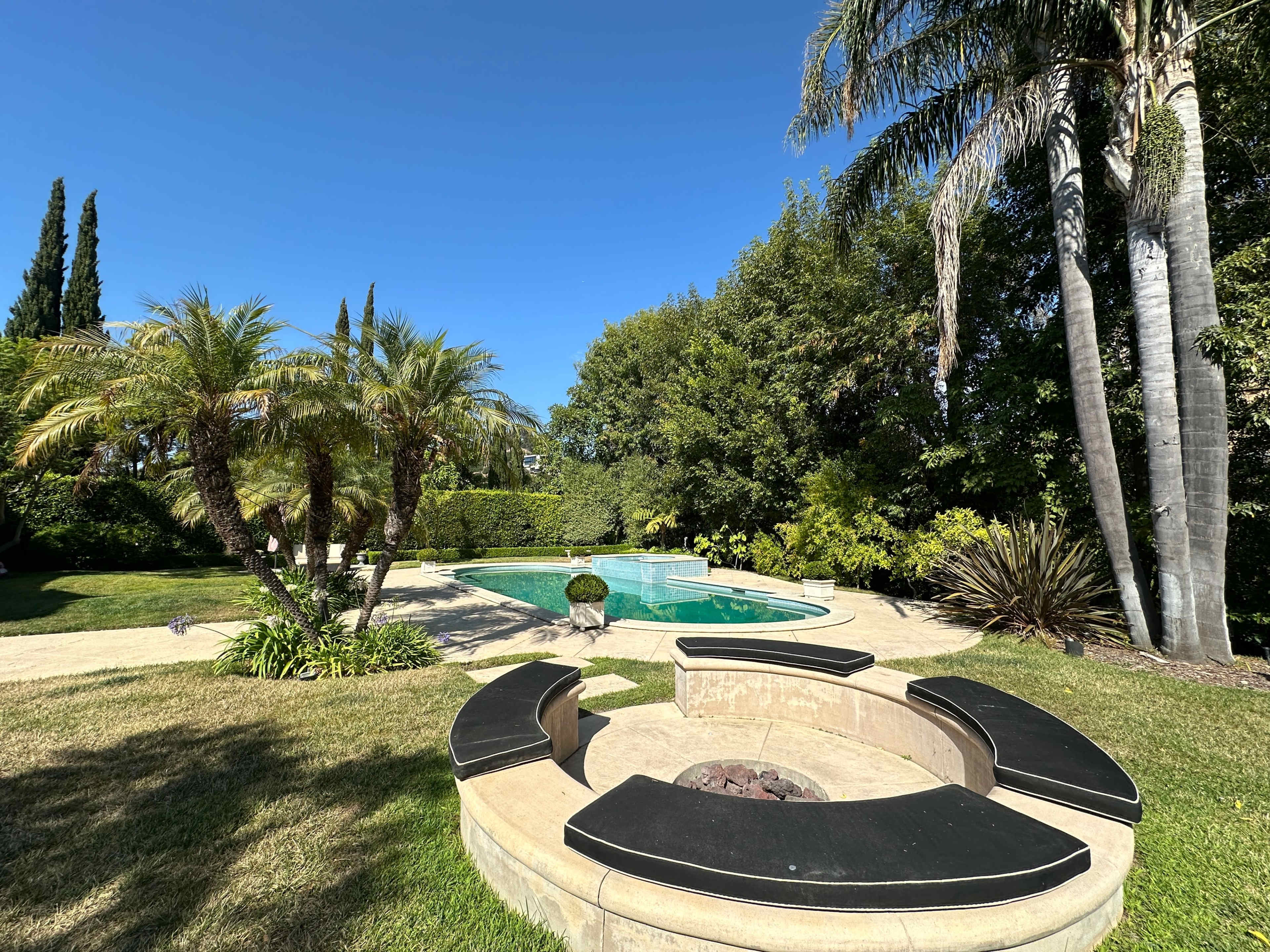 A landscaped backyard featuring a swimming pool surrounded by palm trees and a circular seating area with a fire pit.