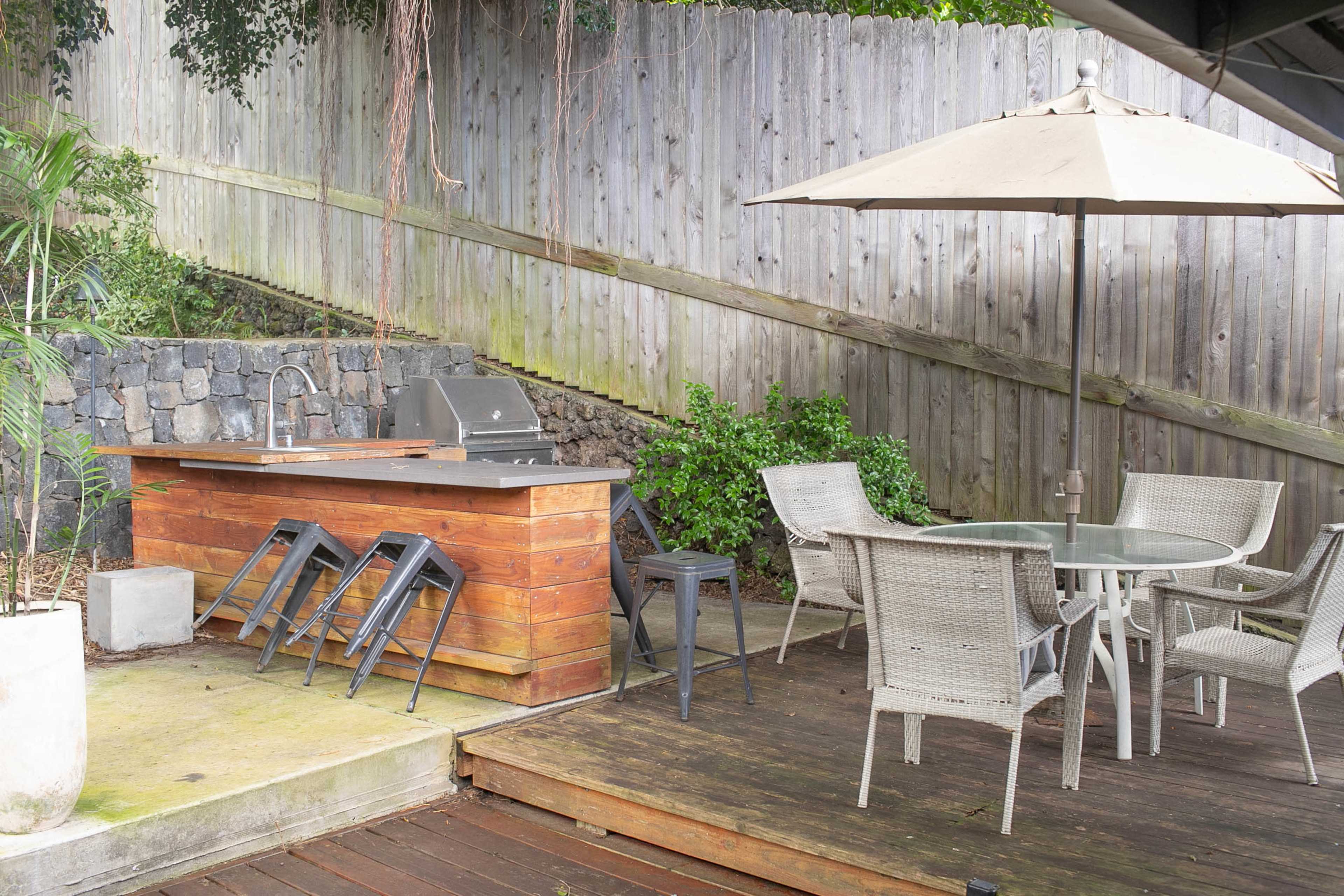 An outdoor patio area with a wooden deck, a modern barbecue grill, a table with chairs, and a large umbrella, surrounded by a wooden fence and greenery.