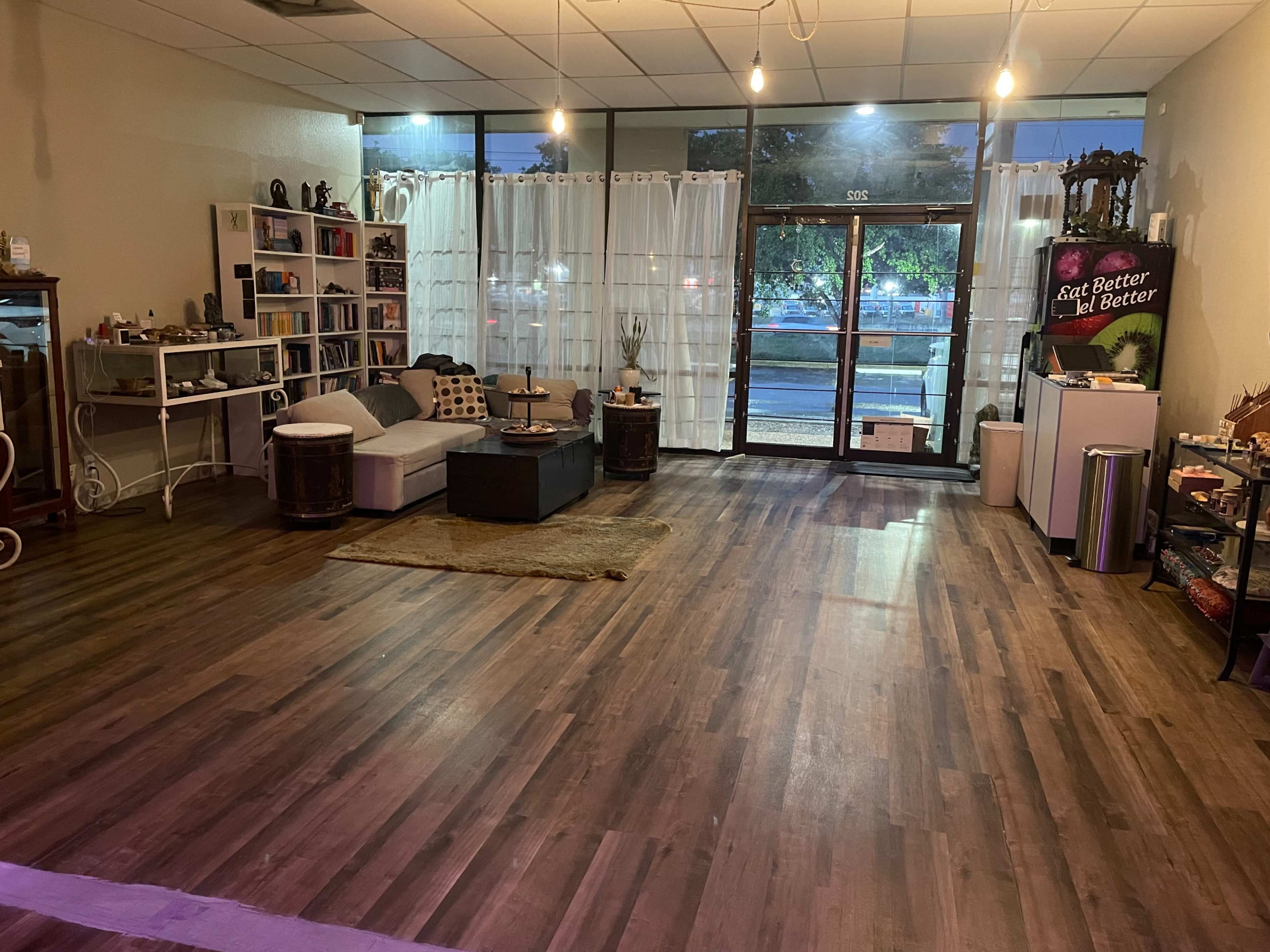 The image shows a spacious, well-lit interior of a room featuring hardwood floors, a sofa with decorative cushions, shelves with books, and a large glass entrance.