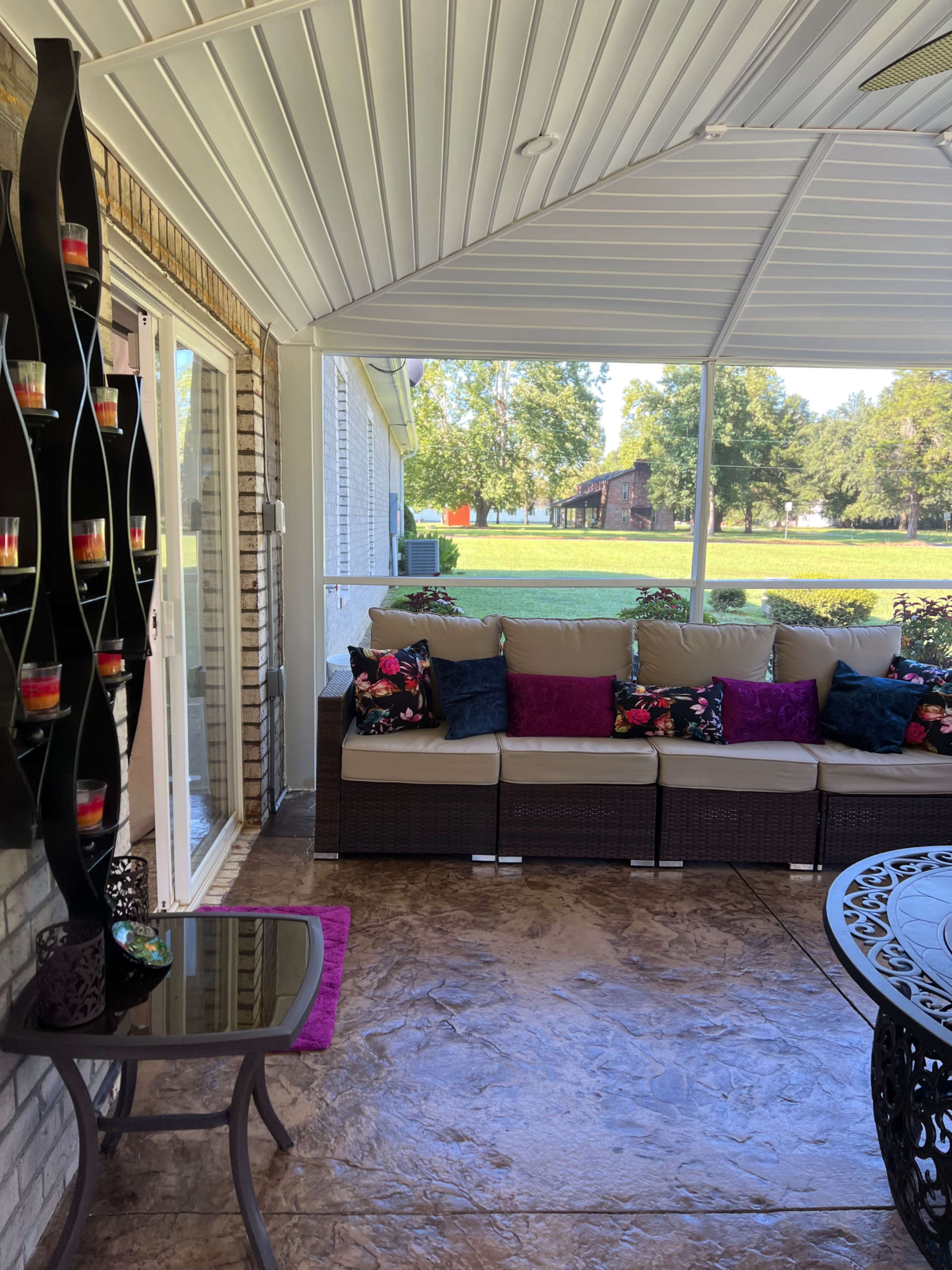 A covered porch with a sofa, decorative pillows, and a table, overlooking a grassy area and trees.