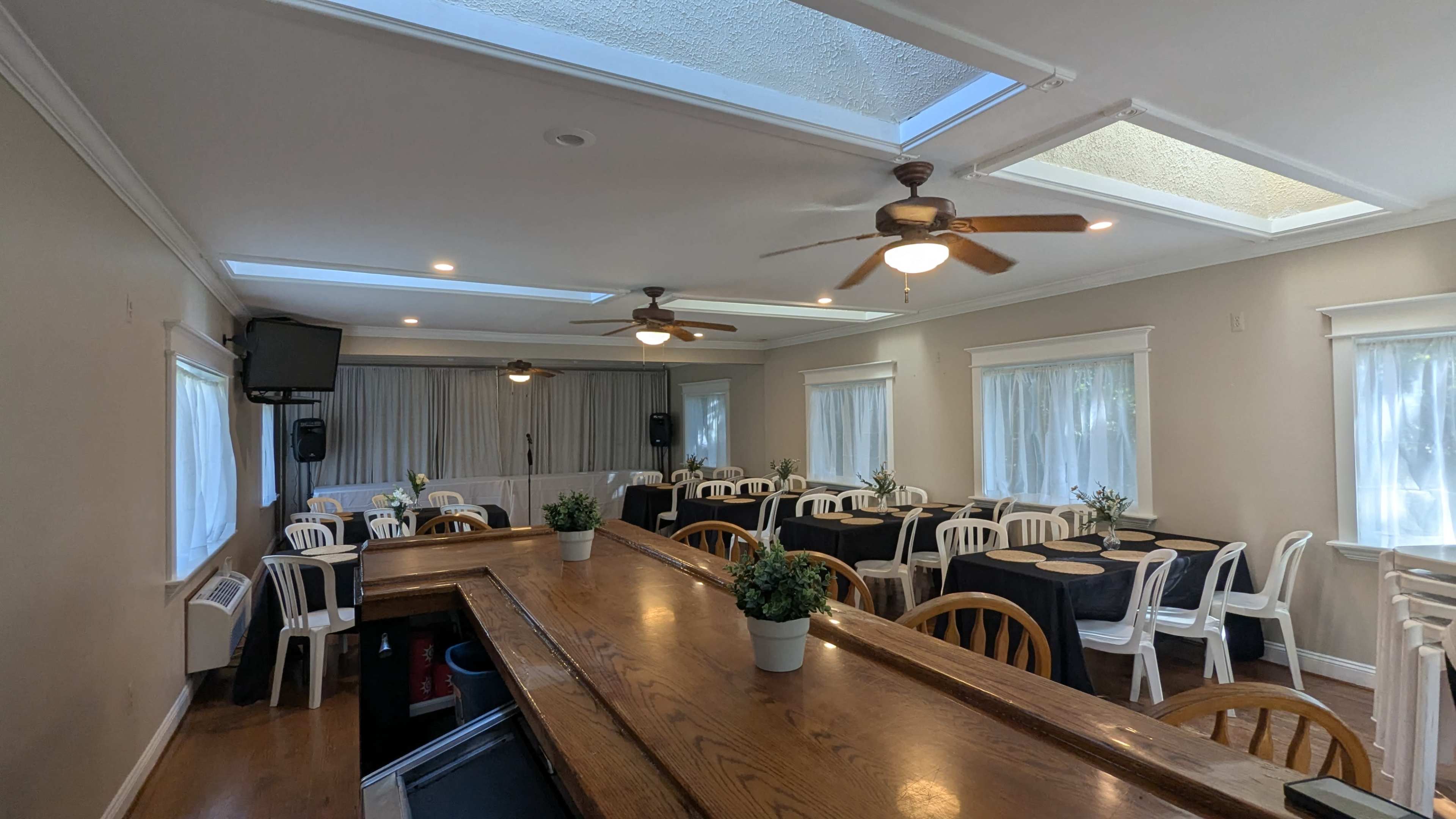 The room features a wooden bar in the foreground and several tables with chairs arranged for an event, with a stage area and windows allowing natural light.